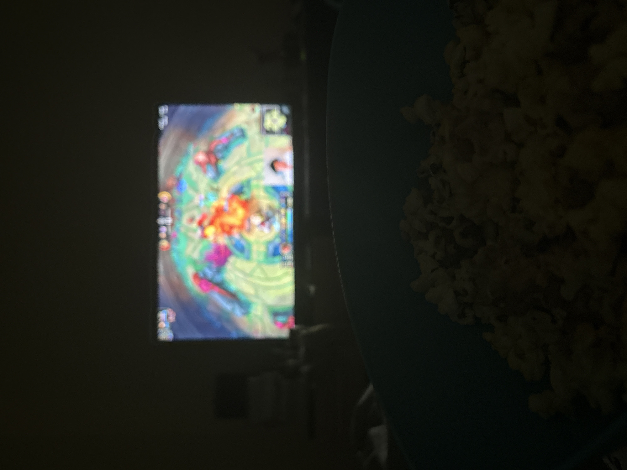 The image depicts a dark room with a bowl of popcorn in the foreground, partially obscuring the view. In the background, a brightly lit screen displays what appears to be a video game. The game features a top-down perspective with colorful visuals and character icons.