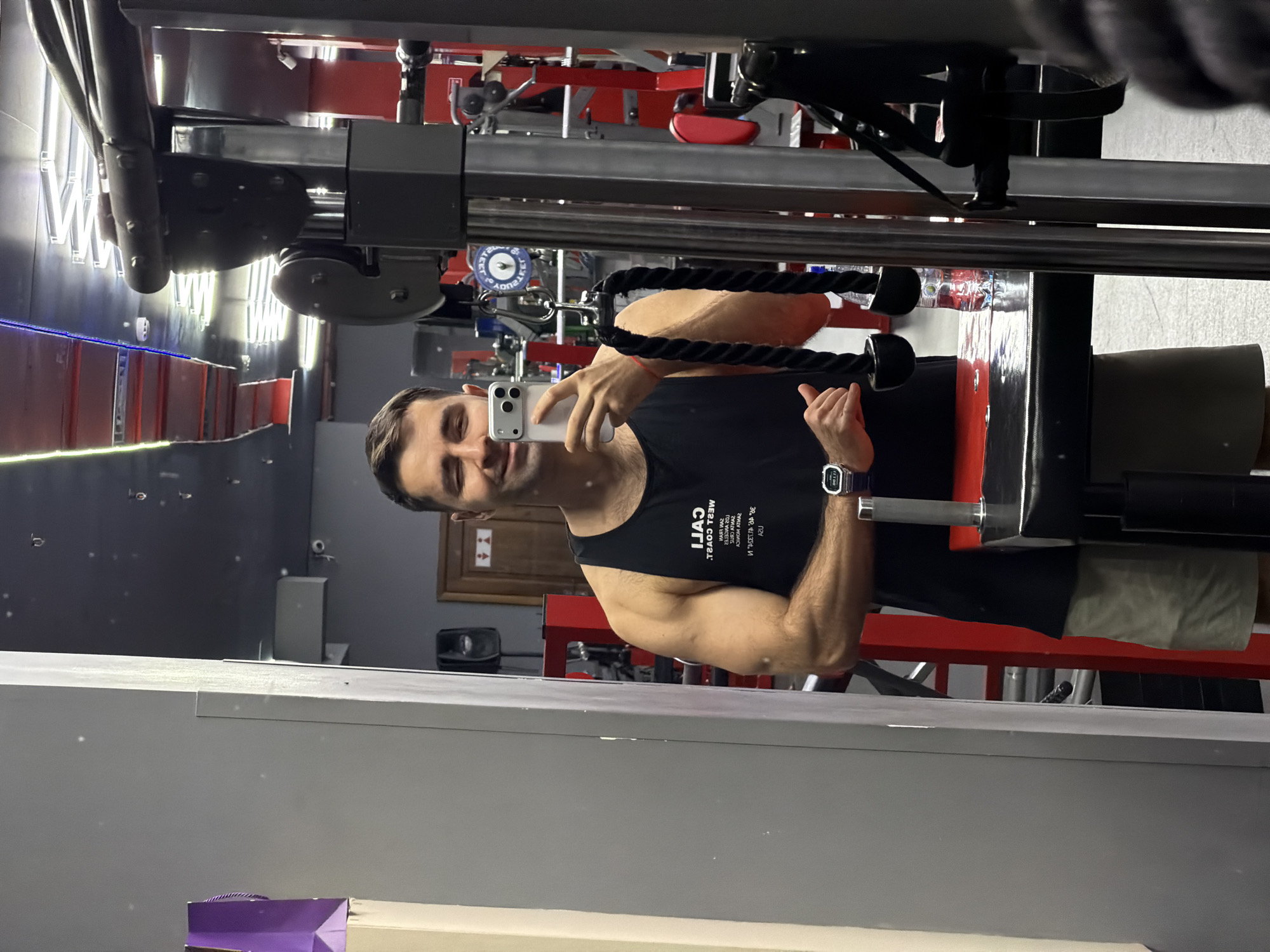 A man is taking a selfie in a gym. He is using a phone to capture the image, holding it up in front of him. He is facing the camera, wearing a black tank top and shorts. The background reveals gym equipment, including a cable machine. The scene also includes a mirror in which the man is taking his picture. No location specifics are available.