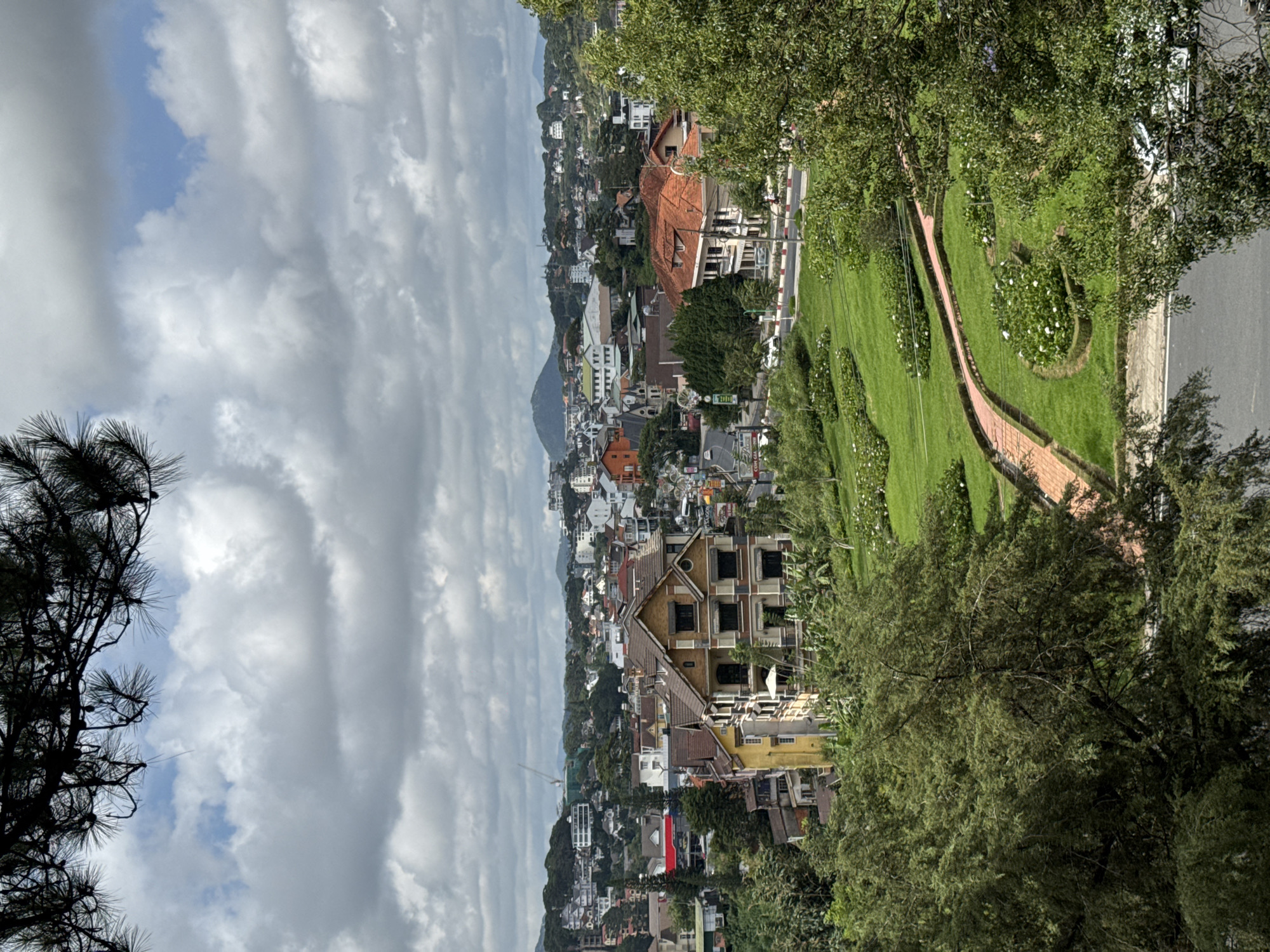 This is a high-angle, outdoor, daytime, full shot of a city landscape in Da Lat, Vietnam. The foreground features lush green trees and manicured lawns with a brick pathway winding through flower beds. Beyond the park, a cityscape unfolds with various residential and commercial buildings, many featuring tiled roofs. A prominent yellow building with brown trim stands out among the others. In the distance, rolling hills are covered in trees and more buildings, with a larger, rounded mountain visible on the horizon under a cloudy sky. The scene is peaceful and serene.