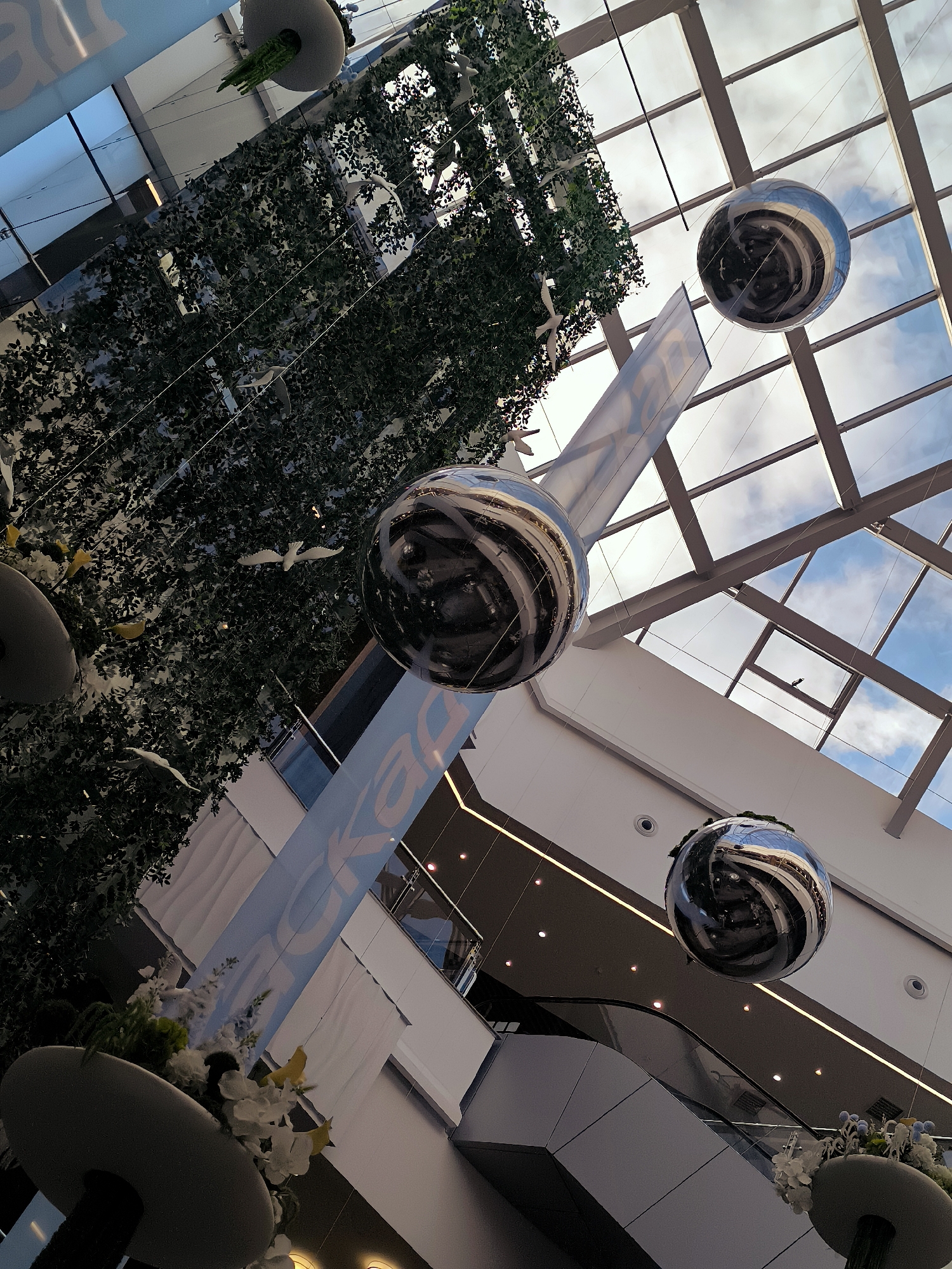 The image depicts the interior of a modern building, possibly a shopping mall or a public space, with a distinctive ceiling structure. The ceiling is a grid of metal beams with glass panels, allowing natural light to filter through, revealing a cloudy sky. Suspended from this ceiling are several large, spherical, highly reflective ornaments that mirror the surroundings, including the sky and the building's interior.
One prominent element is a large, artificial tree or plant installation, dense with dark green foliage, suspended in the upper left portion of the frame. Interspersed with the foliage are small, white, bird-like ornaments, adding a decorative touch.
A large, light blue banner with white lettering is visible, angled diagonally across the center of the image. The lettering is partially obscured but appears to be a brand name or a slogan.
On the lower levels of the building, architectural features such as escalators and balconies are visible. There are also decorative elements like planters with white and yellow flowers. The overall aesthetic suggests a contemporary design with an emphasis on natural light and decorative installations.
While the prompt mentions "the city of Qarağandı city administration, Kazakhstan," there are no direct visual cues in the image that specifically confirm this location. The architecture and decor appear generic to modern commercial or public buildings found in many urban environments. The scene is devoid of any visible individuals.