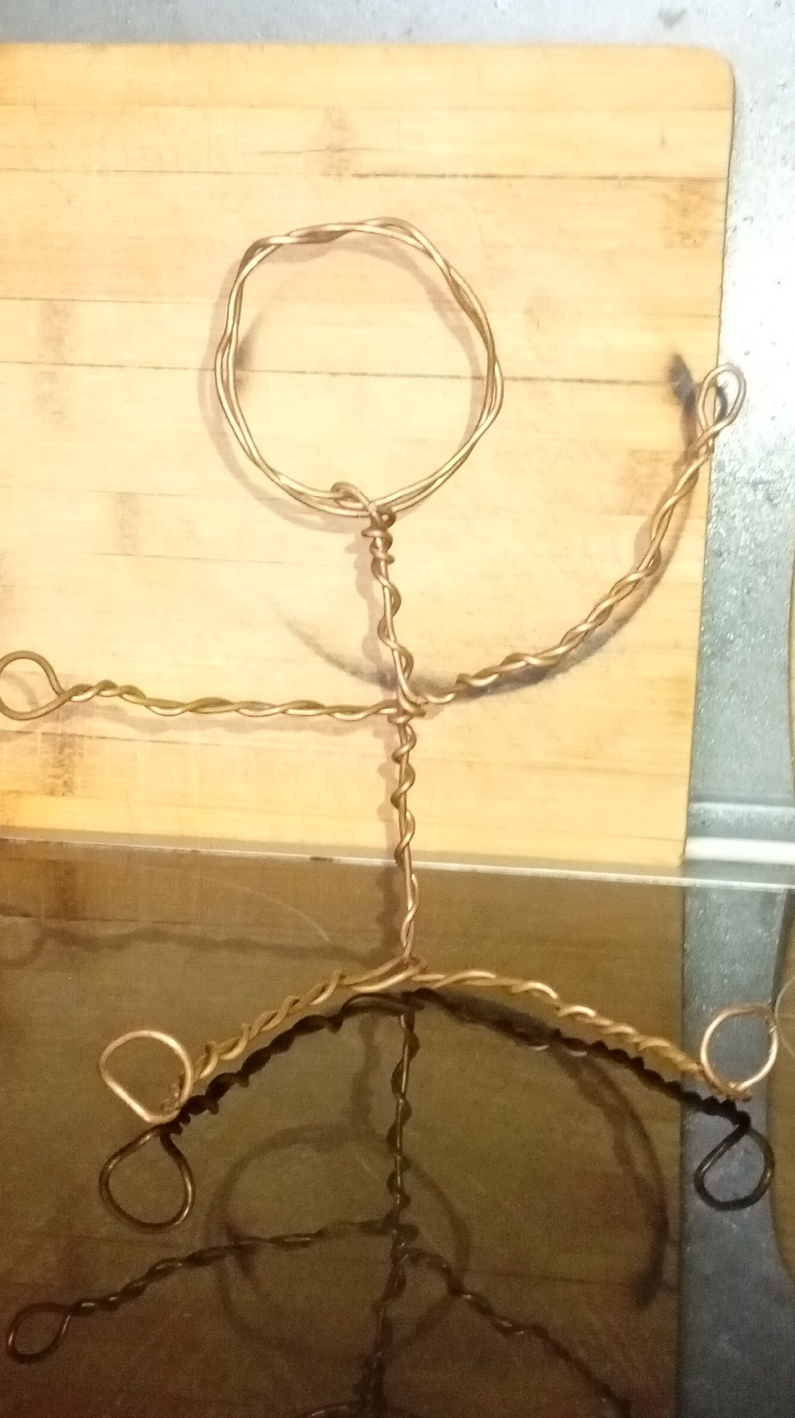 The image shows a wire sculpture of a person on a wooden surface. The sculpture is made of copper-colored wire and depicts a head, torso, arms, and legs. The head is a circular loop, and the torso is a twisted wire forming a straight line. The arms extend outwards from the torso, with one arm bent at the elbow and the other straight. The legs are bent at the knees, forming a seated or crouching position. The sculpture is placed on a wooden cutting board, which is resting on a reflective surface, possibly a table or countertop. The lighting in the image is warm and casts shadows on the wooden surface and the reflective surface. The background is blurred, but appears to be a kitchen environment. The overall impression is of a handmade craft or art piece. There are no people or animals in the image. There is no indication of the location in Karviná, Czechia.