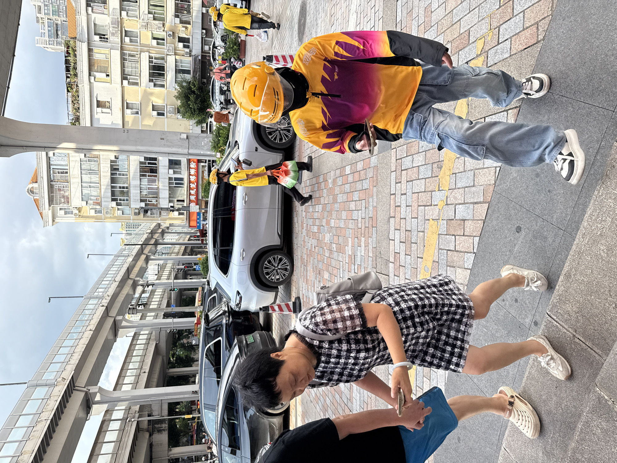 The image depicts a street scene in Haizhu District, China, under an overcast sky. In the foreground, a person wearing a vibrant yellow and purple long-sleeved shirt, blue denim cargo pants, a yellow helmet with a visor, and black athletic shoes is holding a mobile phone. To their left, a woman in a black and white checkered dress and a gray backpack is also looking at her mobile phone while walking. A person in blue shorts and tan clogs is walking ahead of her.
In the background, several vehicles are visible on the street, including a white SUV, a black SUV, and a black sedan. Delivery personnel in yellow shirts and helmets are also present, with one carrying a bag of groceries. Overhead, an elevated highway structure with supporting pillars dominates the upper portion of the image, with buildings lining the street on the right side. The ground is paved with brick and concrete.