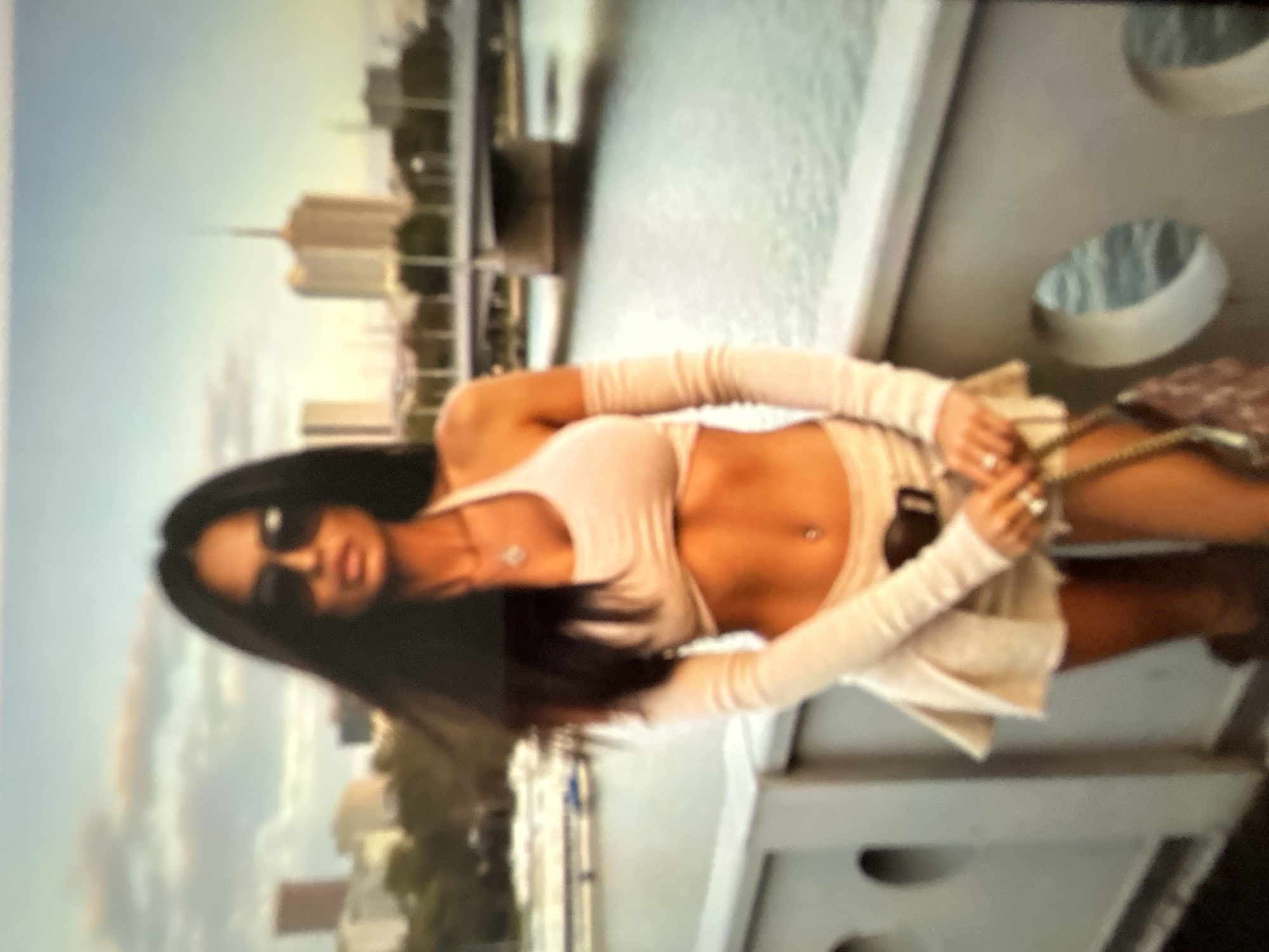 This media file shows a woman with dark hair and sunglasses posing outdoors. She is wearing a light-colored crop top with long sleeves and a matching skirt or shorts. Her midriff is exposed, and she has a belly button piercing. She is holding a purse with a chain strap. The background features a cityscape with buildings and a body of water, possibly a river or bay, suggesting an urban waterfront setting. The lighting indicates it is daytime, likely late afternoon or early evening given the warm tones in the sky. The overall impression is one of a casual, fashion-conscious pose in a scenic location.