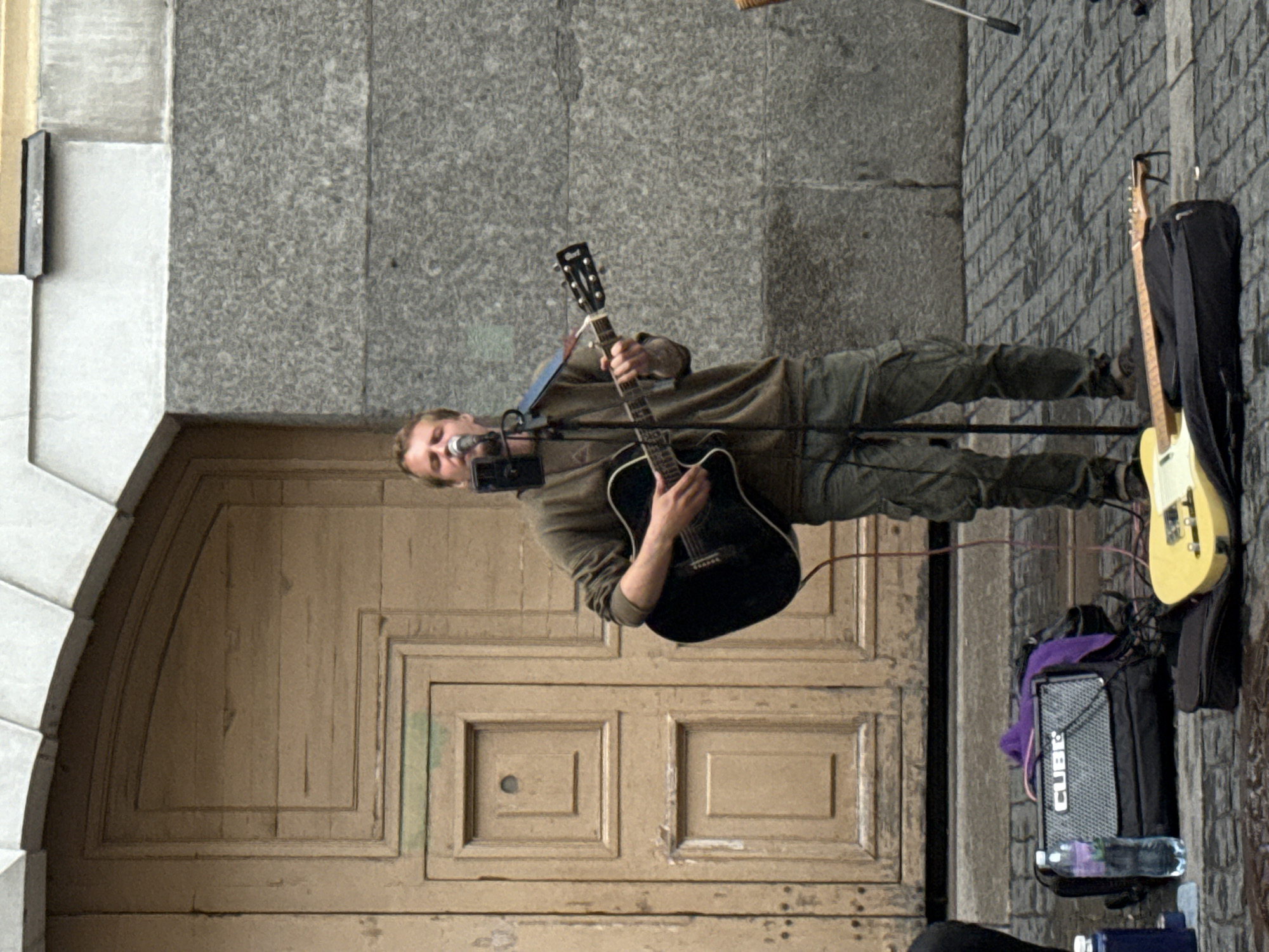 A male street musician is standing and playing an acoustic guitar while singing into a microphone. He is wearing a long-sleeved olive green shirt and camouflage pants. The background consists of a large wooden door with decorative paneling and a gray stone building. In the foreground, there is a guitar amplifier, a water bottle, a purple cloth, and a yellow electric guitar lying on a case. The scene appears to be set on a cobblestone street, possibly in the city of Saint Petersburg, Russia.