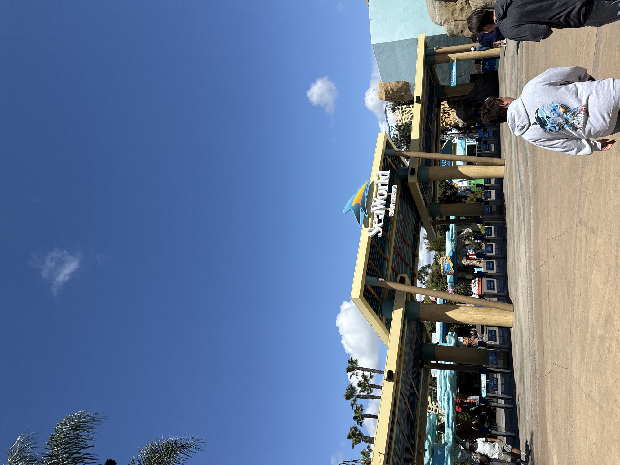 The image captures the entrance of SeaWorld San Diego in San Diego, California, United States. The prominent SeaWorld sign with its iconic blue and yellow wave is visible against a clear, blue sky with scattered clouds. The entrance area consists of several lanes with turnstiles, suggesting a high volume of visitors. People are present, some appearing to be entering the park. The architecture includes a covered structure with columns, possibly for shade or shelter. Palm trees are visible in the background, typical of the Southern California environment. The foreground shows a paved pathway or plaza leading into the park.