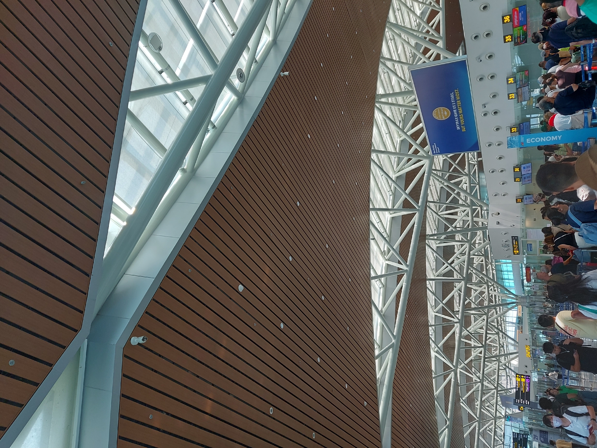 The image depicts the interior of an airport terminal. The ceiling features a modern design with wooden panels and exposed white metal structural elements, interspersed with large glass sections that allow natural light to filter in. Security cameras are visible mounted on the ceiling. Below the ceiling, a large digital billboard displays "Da Nang International Airport" and states "SKYTRAX gave us 5 stars, but yours matter most." Several check-in counters are lined up, numbered 28 through 36, with digital displays indicating flight information. A vertical blue sign reads "ECONOMY." A crowd of diverse individuals, appearing to be travelers, are queuing at the check-in counters. Many are carrying luggage. The overall atmosphere suggests a busy airport environment.