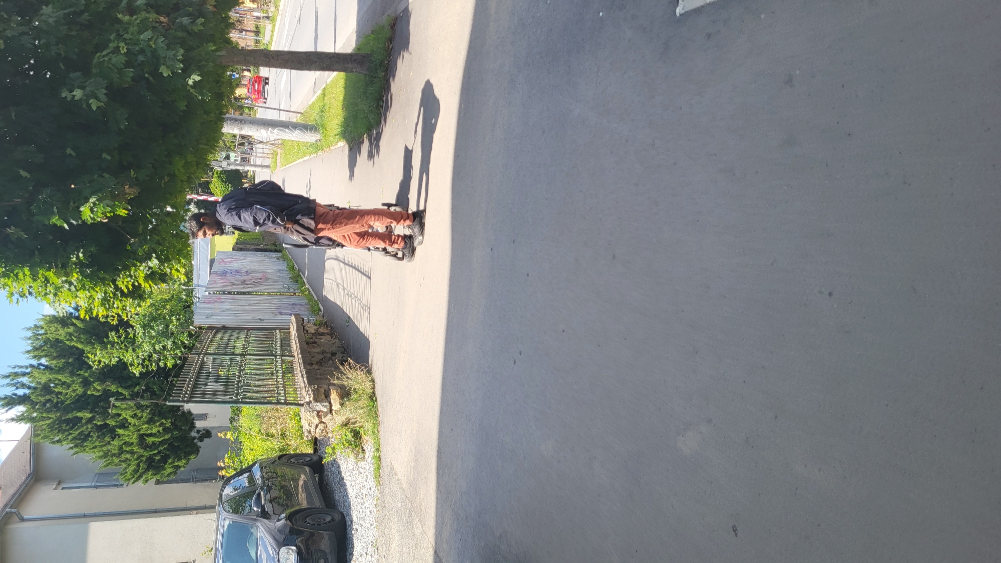 A man, wearing a dark jacket and orange pants, stands on a sidewalk in Prešov, Slovakia, pushing a stroller. He is facing away from the camera, looking towards a street with a red car driving in the distance. The sidewalk is paved with concrete and bordered by a metal fence and a row of trees. To the left, a black car is parked on a gravel surface next to a building. The scene is illuminated by sunlight, casting shadows on the ground.