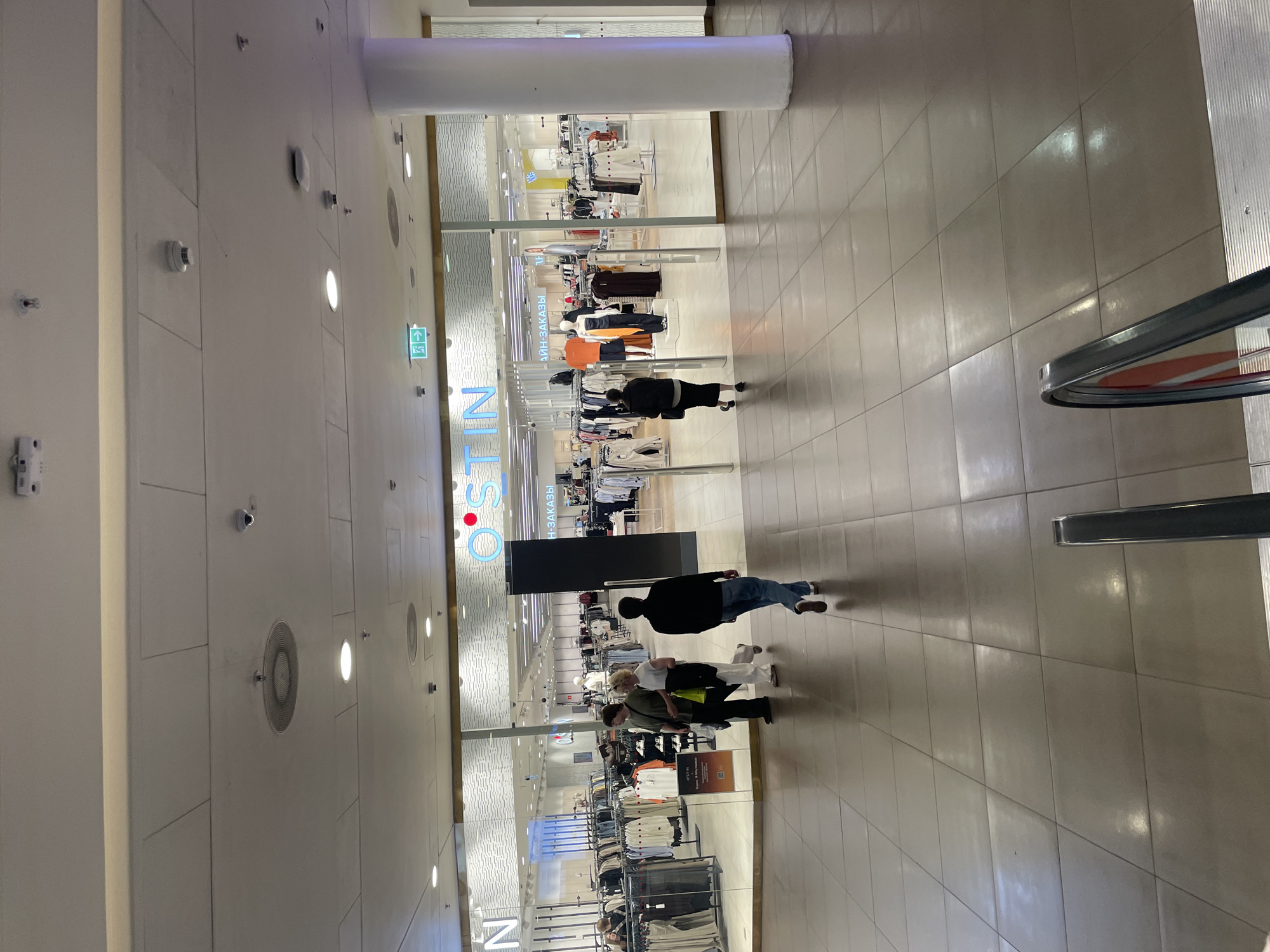 The image captures an indoor scene within a commercial building, likely a shopping mall in Saint Petersburg, Russia. The architectural features include a high ceiling with recessed lighting and ventilation systems. A prominent store sign, readable as "OSTIN," is visible, indicating the presence of a clothing retailer. Mannequins display various clothing items inside the store, and shoppers are seen browsing or walking through the open store entrance. The mall floor is tiled, and an escalator is present on the right side of the image. Several individuals are in view, mostly casually dressed, and they appear to be shopping or passing through the mall area.