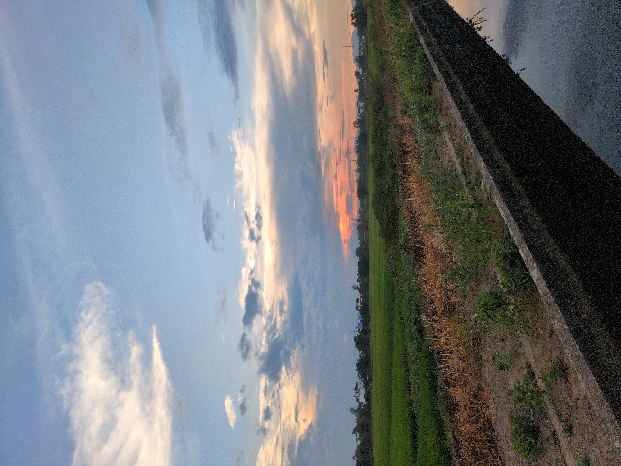 The image depicts a rural landscape at dusk, likely in the vicinity of Hoi An, Vietnam. The sky is filled with a mix of blue, white, and orange clouds, indicating the sun is setting. A bright orange glow is visible near the horizon, suggesting the sun is just below it. In the foreground, a concrete canal runs diagonally across the frame, reflecting the dark sky and surrounding vegetation. The banks of the canal are lined with sparse green plants and dry, golden grass. Beyond the canal, a vibrant green rice paddy stretches across the middle ground, interspersed with patches of dry earth and more vegetation. In the distance, a line of trees separates the fields from a hazy horizon where a few structures and distant mountains can be faintly discerned. The scene is tranquil and natural, with no people or animals visible. The presence of rice paddies and the style of rural housing in the distance are consistent with the agricultural outskirts of Hoi An.