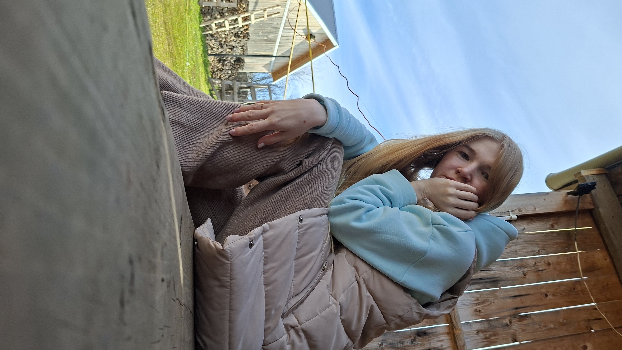 A young woman with long, straight, reddish-blonde hair is captured in an outdoor setting, looking directly at the viewer from a low, slightly upward angle. She is dressed in a light blue long-sleeved top, brown ribbed pants, and a light beige sleeveless puffer vest. Her left hand, featuring a golden ring on her ring finger and light-colored nail polish, rests on her bent knee. Her right hand is brought up towards her chin, partially obscuring her mouth. Her expression is neutral and direct.
She appears to be sitting or leaning against a wooden structure that forms a significant part of the foreground and mid-ground on the left and right, possibly a wooden bench, deck, or a shed wall. In the background, under a clear, bright blue sky with faint wisps of clouds, a rural scene unfolds. It includes a grassy area, a large pile of stacked firewood, and a rustic wooden building, likely a barn or shed, with a light-colored roof. A black cable and a light-colored pipe are also visible against the wooden structure on the right side of the frame. The scene depicts a calm and bright day in the countryside, likely in Большой Букор, Russia. There is no visible text in the image.