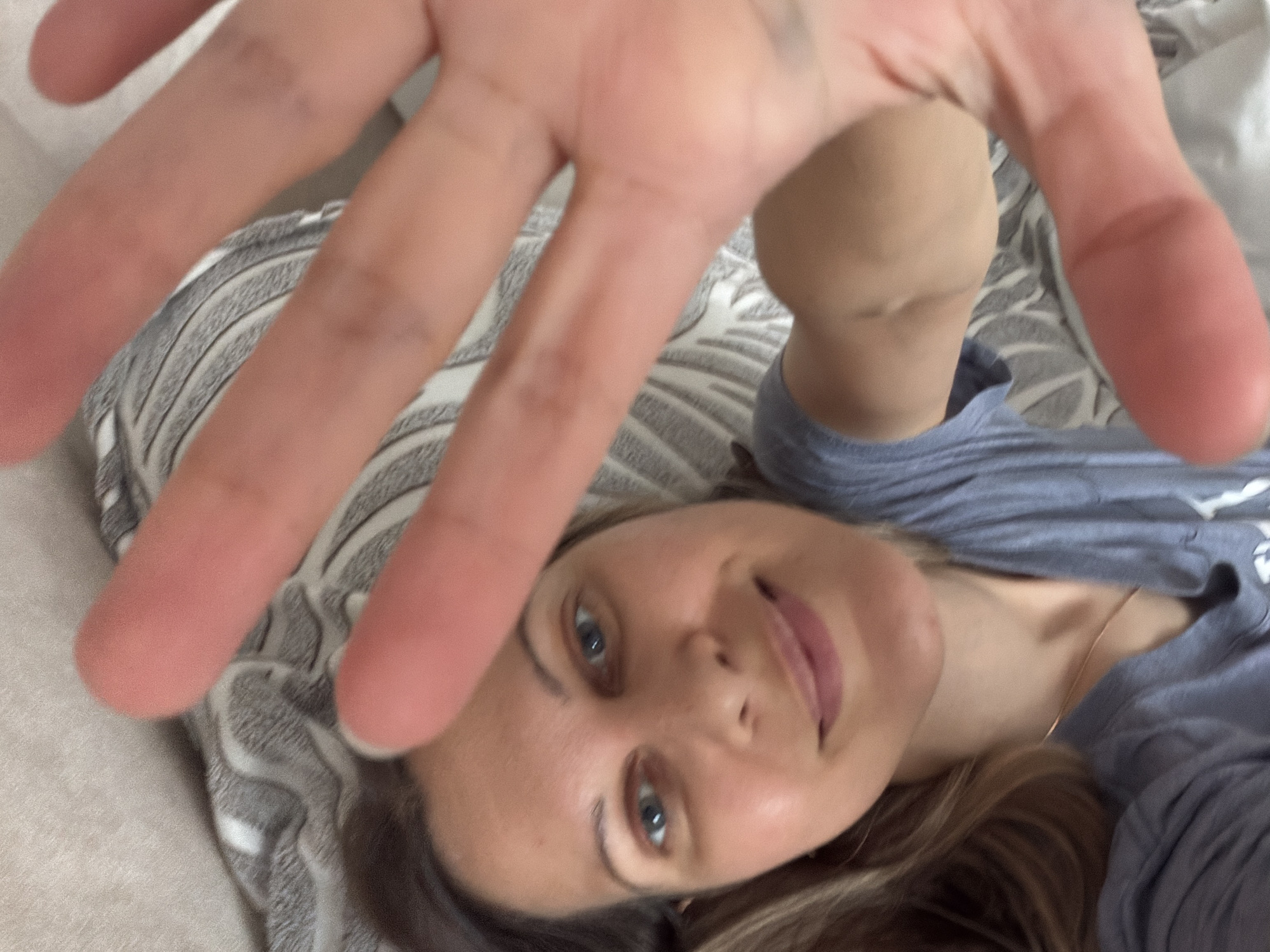 A woman with blonde hair and blue eyes is lying down and holding her hand up to the camera, obscuring part of the image. She is wearing a grey t-shirt and a thin gold necklace. She is on a bed with a striped blanket. The image is blurry, and it is not possible to determine the location.