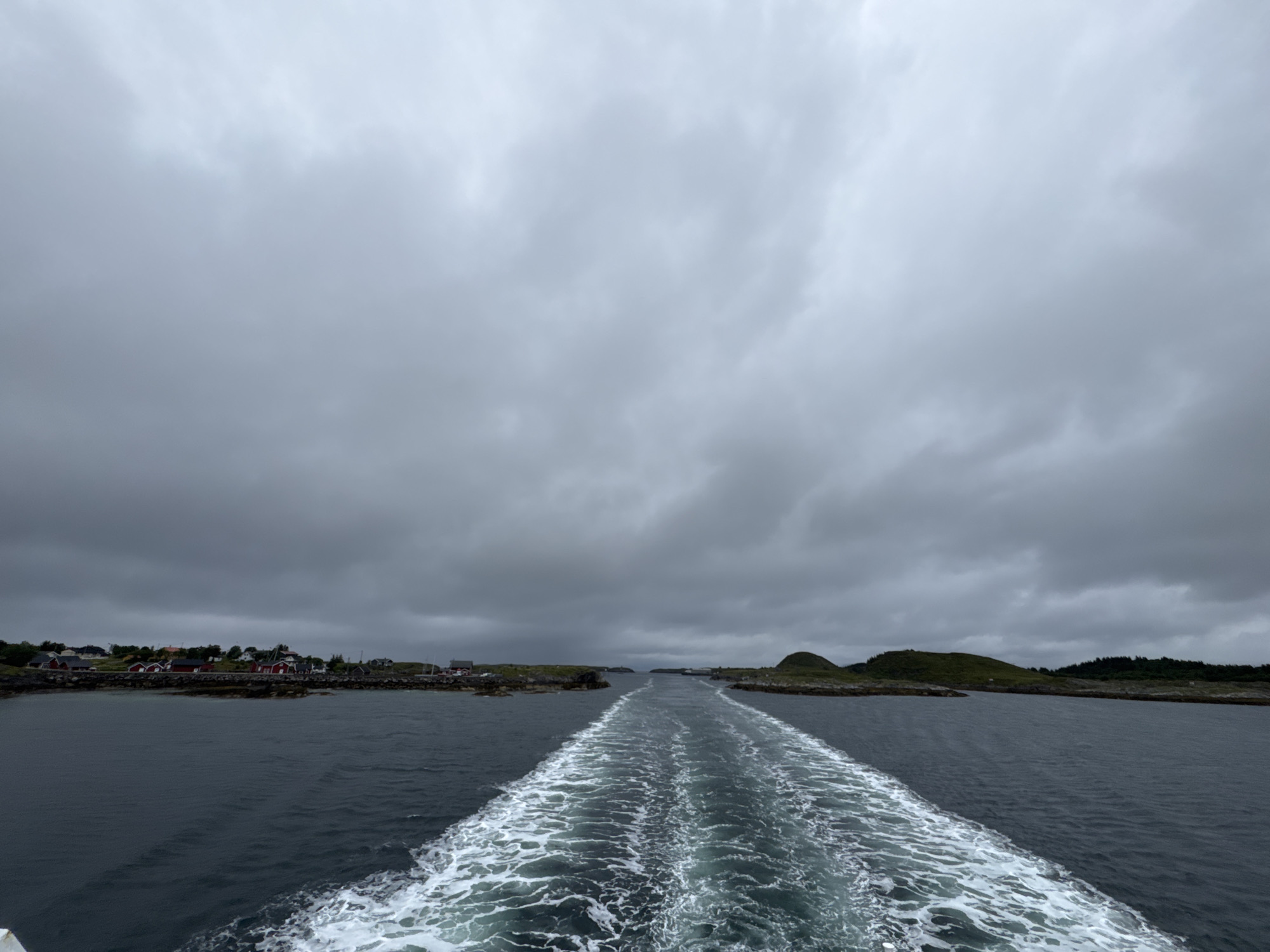 The image depicts a view from a boat moving away from a settlement on Sleneset, Norway. The water is dark and choppy, with a prominent white wake trailing behind the boat. The shore, visible on the left, is lined with red-roofed buildings and green, rocky terrain. The sky is overcast with heavy grey clouds. The scene suggests a journey through a fjord or coastal area under a cloudy, possibly cool, day. There are no people visible in the image.