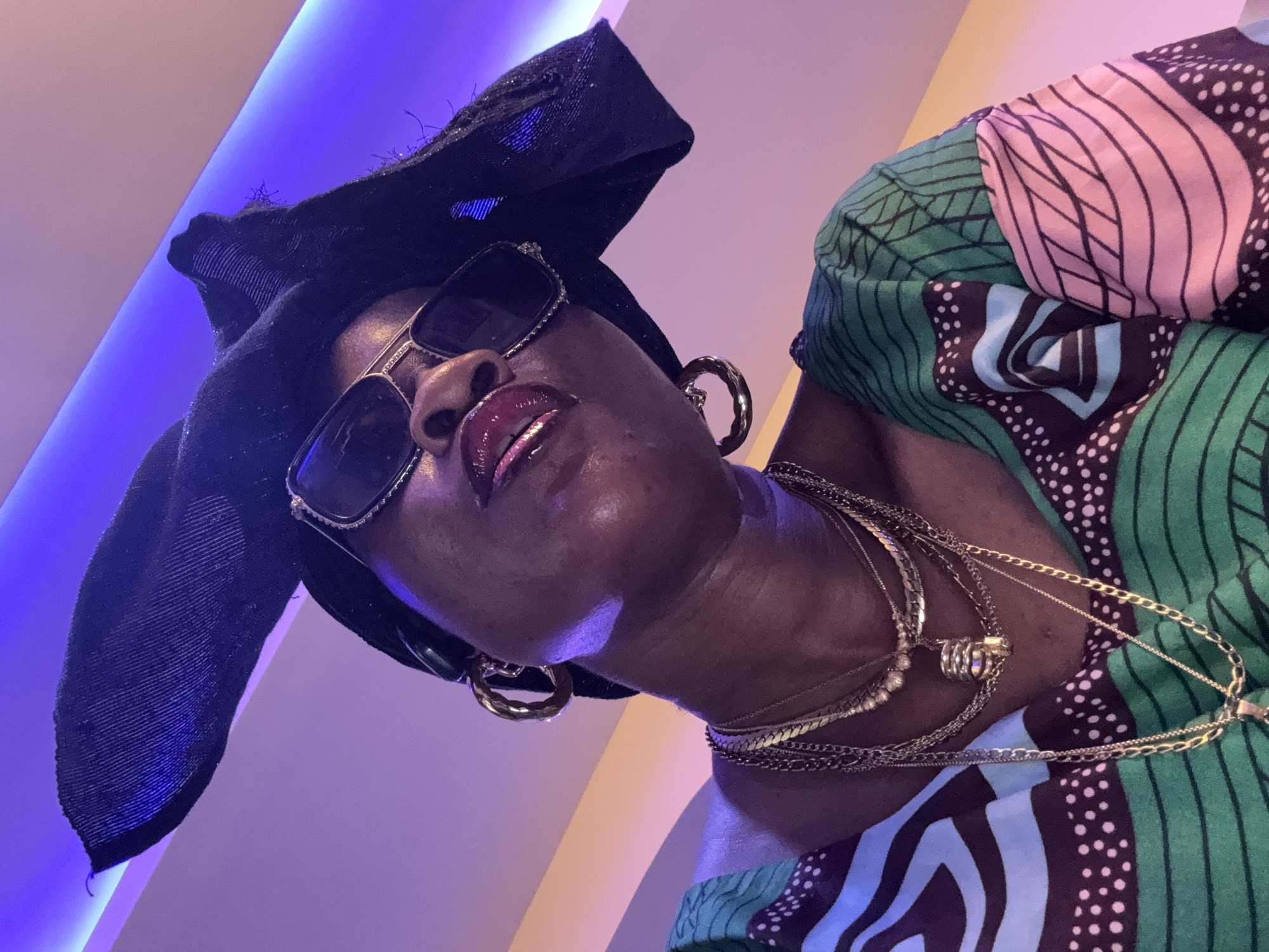 A low-angle, medium shot captures a woman from the chest up, wearing a black headwrap and large sunglasses. Her head is tilted upwards, and she is looking directly at the camera. She has dark skin, full lips with glossy lipstick, and prominent cheekbones. She is adorned with multiple gold necklaces and large gold hoop earrings. Her green and patterned top features geometric shapes in pink, blue, and white. The background consists of a wall illuminated by purple and orange lighting. The scene is indoors, the location within Ajara, Nigeria, is not specified.