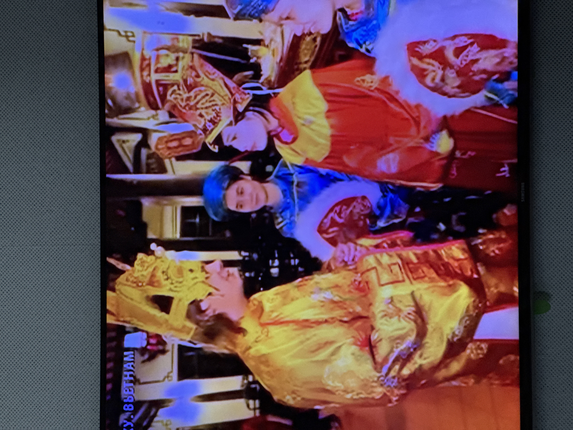 The image shows four people in ornate, traditional clothing, likely during a performance or ceremony. The person on the far left is wearing a golden robe and a tall, elaborate golden headdress with dragon motifs. To their right, a person in a blue robe and a conical blue hat is holding a fan. Next to them, a person in a red and yellow robe with a similar conical headdress is also holding a fan. On the far right, a person in a blue garment and a beaded headdress is partially visible. The background suggests an indoor setting with architectural details and what appears to be a stage or decorated area. Text in the top left corner of the image reads "КУ. ВЬЕТНАМ", which translates to "Republic of Vietnam" in Russian. This strongly indicates the scene is related to Vietnam, not Oskemen, Kazakhstan.