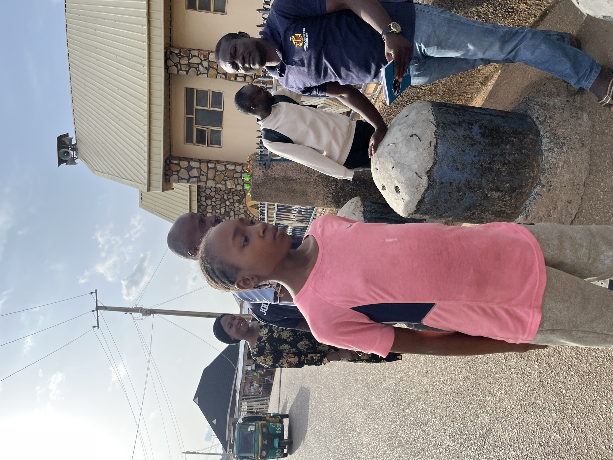 The image shows a group of people standing on a street in Jos, Nigeria. In the foreground, a young girl wearing a pink shirt and grey trousers is looking to the side. Behind her, there are several adults, including a woman in a floral print shirt and a man in a blue polo shirt with an emblem. The man is holding a book or notebook. Another man is wearing a white sweater with black accents. They appear to be standing near a large stone pillar or marker. The background features a building with a stone facade and a metal roof. A speaker is mounted on the roof. A motorized tricycle is parked on the side of the street. Overhead, there is a utility pole with power lines. The scene appears to be outdoors during daylight hours, with a clear sky above.