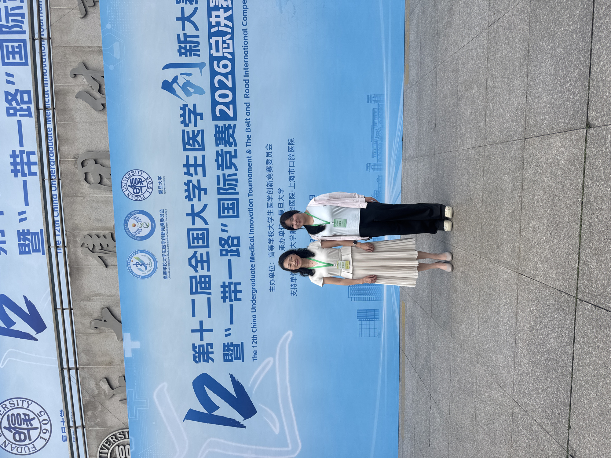 Two women stand in front of a large blue banner with Chinese text and logos. The banner is likely for an event, as indicated by the text which translates to "The 12th China Undergraduate Medical Innovation Tournament & The Belt and Road International Competition 2026 Finals." The woman on the left is wearing a cream-colored pleated dress and heels, while the woman on the right is wearing a white t-shirt, a pink cardigan, and black pants with white shoes. Both women are wearing lanyards with identification badges. The banner features logos for universities and organizations, including "Fudan University." The setting appears to be an outdoor plaza with a tiled floor, and a building facade with architectural lettering is visible in the background. The scene is likely located in Yangpu District, China, given the mention of Fudan University, a prominent institution in that district.