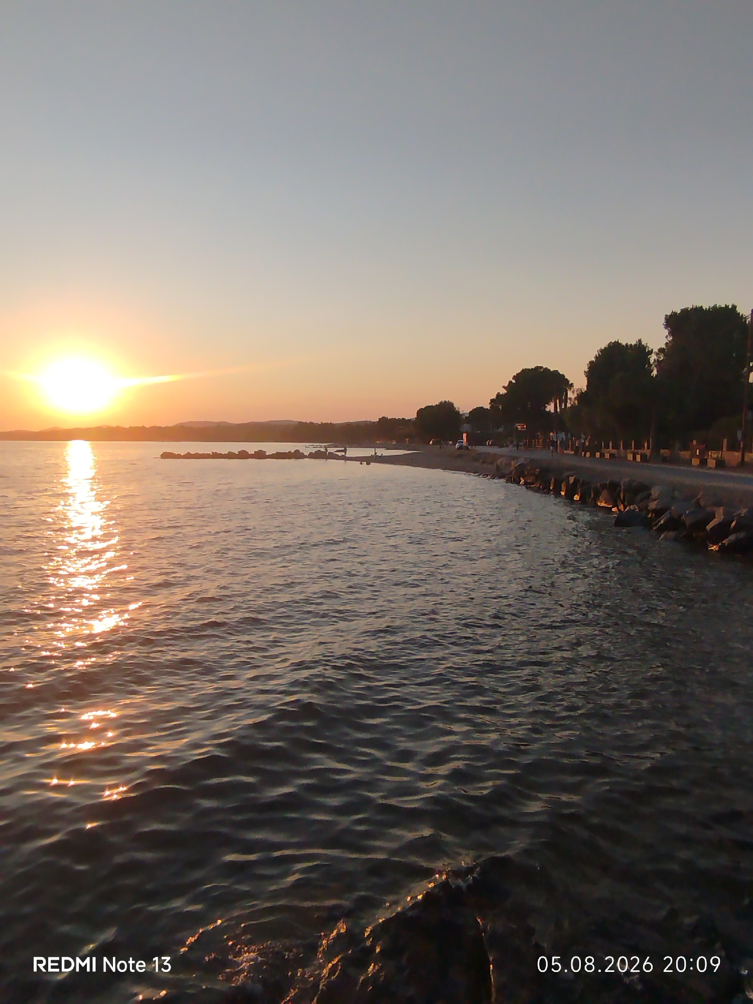 The image depicts a sunset over the sea in Sithonia Municipal Unit, Greece. The sun is low on the horizon, casting a golden glow on the water and the sky. The foreground is dominated by the rippling sea, with a rocky shoreline visible on the right. Further in the distance, there is a paved road with some people and trees lining it. The overall scene is tranquil and serene, characteristic of a Mediterranean coastal setting during sunset.