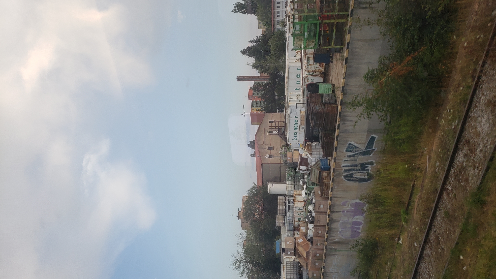 This is an image taken from a moving train, likely on the outskirts of Poprad, Slovakia, as suggested by the industrial and residential buildings visible. The scene depicts a large outdoor storage area filled with various containers, crates, and industrial equipment. In the foreground, train tracks run horizontally across the bottom of the frame, indicating the perspective of the viewer.
A concrete wall runs along the middle ground, adorned with graffiti. To the left, purple lettering is partially visible, while to the right, more prominent dark blue graffiti reads "OHK" in bold, angular letters. Behind this wall, a collection of white and metallic shipping containers are stacked and arranged. Some appear to be industrial tanks, while others are more standard storage units. Among them, there are stacks of wooden crates, metal structures, and various other industrial materials.
In the background, several buildings can be seen. To the left, there is a low-rise, light brown industrial building with a red roof. Further back and to the right, a taller, multi-story building with colorful facade (orange, yellow, green) stands out, alongside other apartment buildings and a tall, narrow brick chimney stack. Trees and foliage are scattered throughout the scene, particularly around the buildings. The sky above is partly cloudy, with a mix of blue sky and white clouds. The overall impression is one of an industrial zone or a logistics hub situated near a residential area.