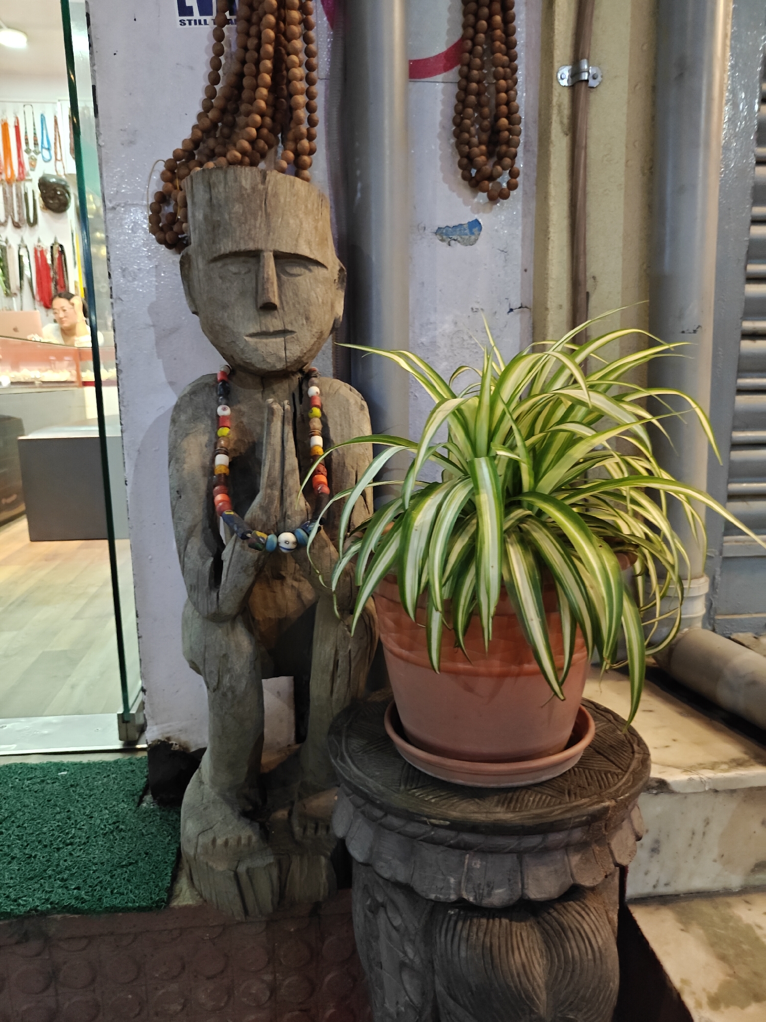 A wooden statue of a seated figure with hands pressed together in prayer is positioned next to a potted spider plant. The statue has a wooden necklace with colorful beads and a string of brown wooden beads hanging from its head. In the background, a store window displays various necklaces and accessories, with a person seen working inside. The scene appears to be outdoors, with a green mat on the ground in front of the statue and a marble step to the right. The overall setting suggests an entrance to a shop or a marketplace in Kathmandu Metropolitan City, Nepal.