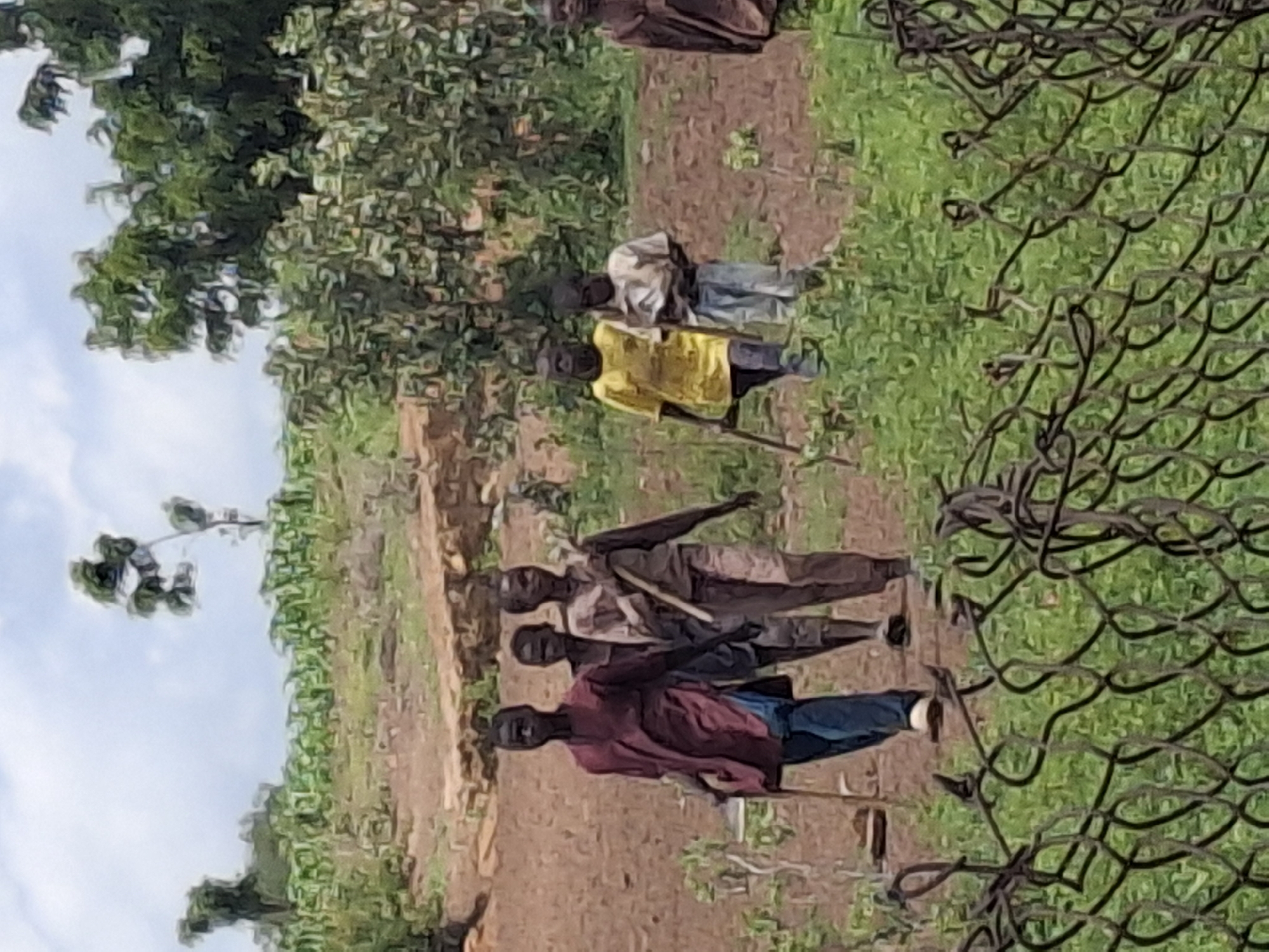 A group of six young men and boys are walking across a dirt path in a rural, outdoor setting. The individuals are dressed in casual, loose-fitting clothing, with some carrying sticks. The environment features sparse vegetation, including bushes and trees, and the ground is mostly bare earth. The sky is partly cloudy. The scene appears to be in a village area, possibly in Lembi, Nigeria.