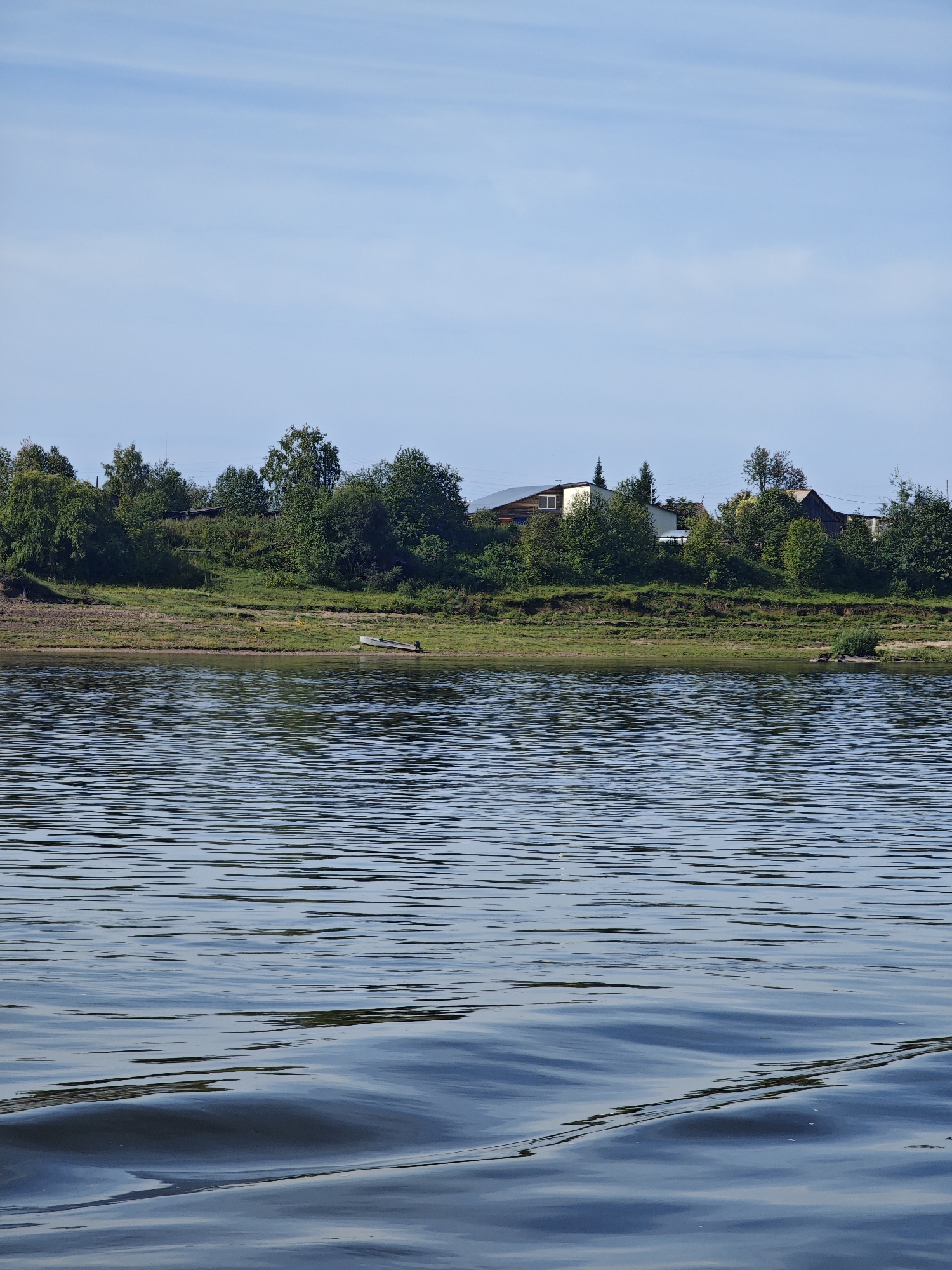 This image depicts a serene river scene, likely in or near the city of Yartsevo, Russia, on a clear day with a partly cloudy blue sky. The foreground is dominated by the reflective surface of a body of water, exhibiting gentle ripples and waves that catch the light, creating a shimmering effect. In the middle ground, a grassy bank lined with trees forms the edge of the river. A small, light-colored rowboat with an outboard motor is pulled ashore on the grassy bank. Beyond the trees, a few houses with distinct rooflines are visible, suggesting a rural or semi-rural settlement. The vegetation is lush and green, indicating a warm season. The overall atmosphere is tranquil and natural.