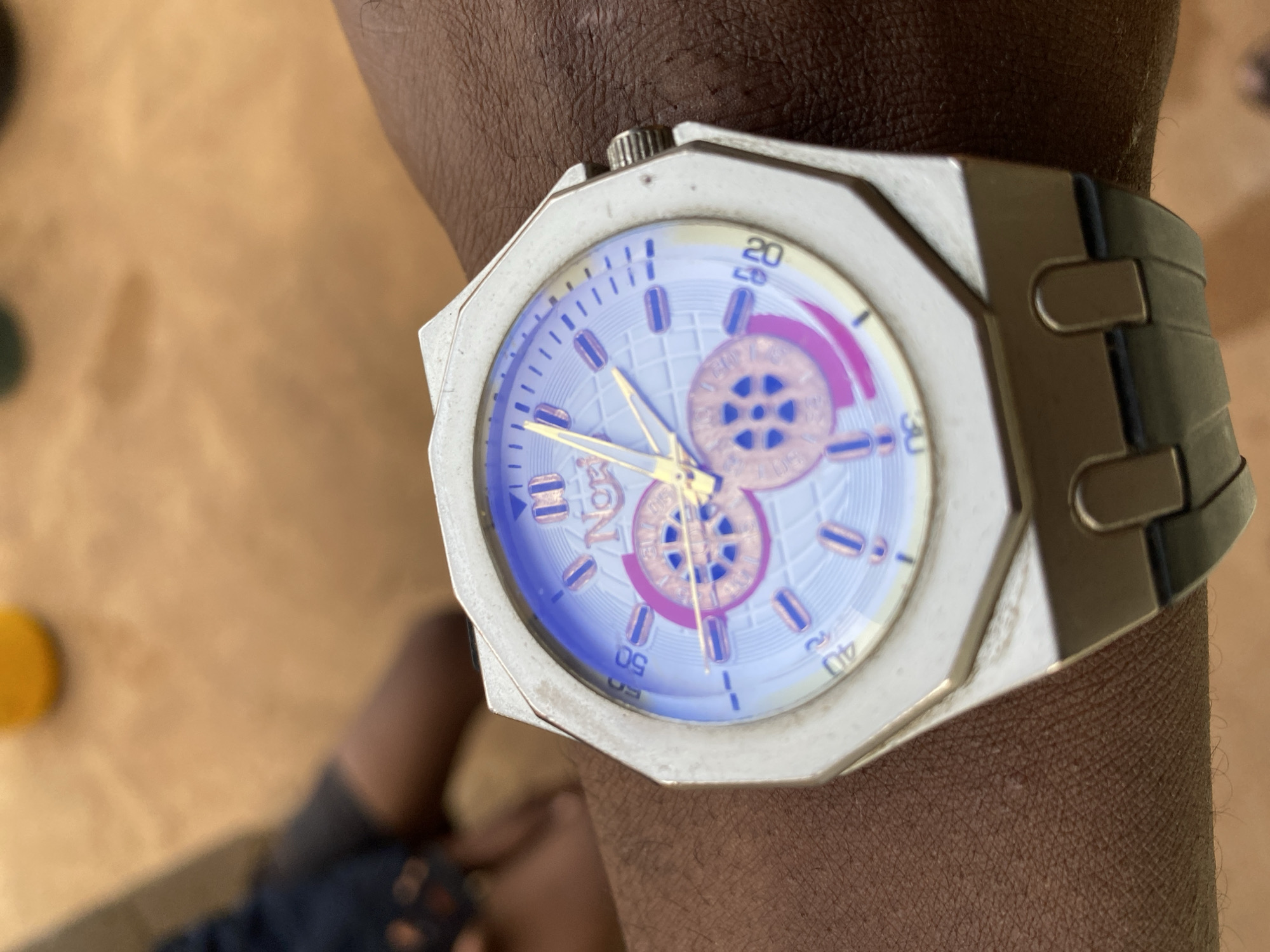 A wristwatch is on a dark-skinned wrist. The watch has a light-colored, octagonal face with a light blue dial, gold hands, and a black band. The watch face has markers at each hour and small circular insets at the top and bottom. The background is blurry, showing a portion of the subject's leg, clothed in blue jeans with a patterned design, and a tan-colored floor. The location is unknown, and there is no verifiable evidence that this image was taken in Bukuru, Nigeria.
