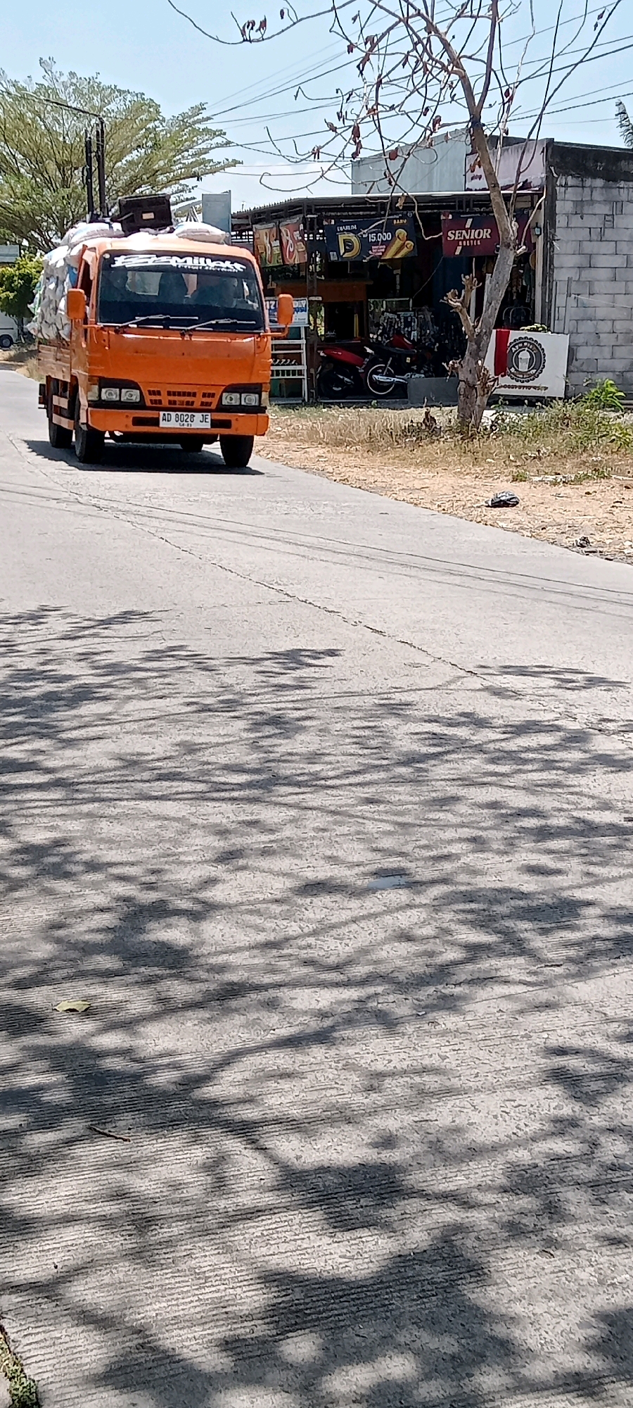 An orange truck with a license plate reading "AD 8028 JE" drives down a paved road in Desa Karangmojo, Indonesia. The truck is loaded with white sacks, some of which appear to be plastic bags. In the background, there are buildings with awnings and signage. One sign reads "SENIOR BOOTIK." There are also motorcycles parked in front of the buildings. Trees with sparse foliage are visible, and their shadows are cast across the road. The sky is blue and clear.