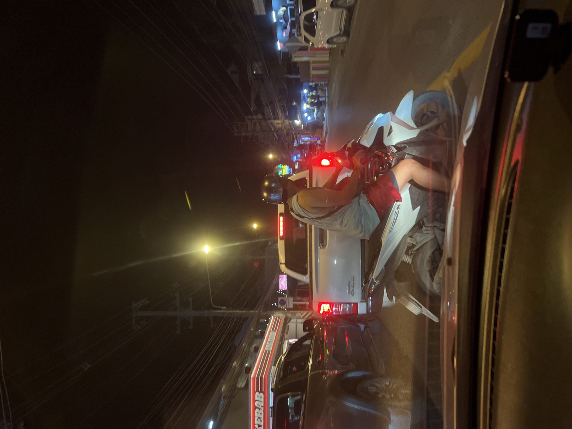 In the city of Ko Samui District, Thailand, at night, a person wearing a helmet and red shorts is riding a white scooter. The scooter is positioned behind a white pickup truck. To the left of the pickup truck, a dark-colored SUV is visible. In the background, there are buildings, streetlights, and power lines. The scene appears to be a street with traffic, possibly a commercial area given the presence of a "KEBAB" sign on a building.