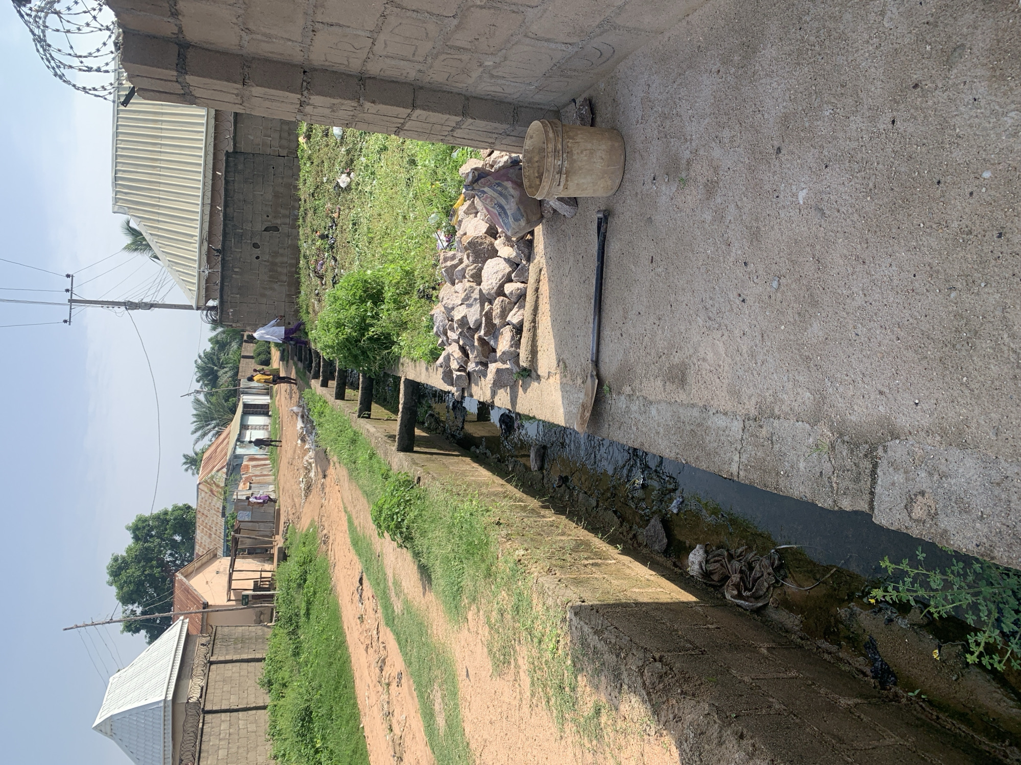 The image depicts an outdoor scene in what appears to be a residential area with a pathway and a drainage ditch. The orientation of the image is rotated 90 degrees clockwise.
The foreground is dominated by a concrete pathway and a narrow, dark drainage ditch running alongside it. The ditch contains some murky water and debris, including what looks like a piece of cloth. To the left of the pathway, a dirt track with sparse green grass is visible. Beyond the dirt track, several buildings with corrugated metal roofs and brick walls are scattered. There are also trees and vegetation in the background.
In the middle ground, on the pathway near a pile of rocks and a bucket, a few people are visible. One person, wearing white clothing, is standing. Two other figures, appearing to be children or younger individuals, are also present. They are walking along the pathway.
In the background, more residential structures are visible, some with tin roofs. A power line pole with wires stretches across the scene. The sky is bright, suggesting it is daytime, and the weather appears to be clear. The overall setting suggests a somewhat undeveloped or rural area.
Notable details include the construction materials like bricks and corrugated metal, the presence of a drainage ditch which may indicate sanitation challenges, and the natural elements like grass and trees. The scene conveys a sense of everyday life in this location. There is no visible text in the image.