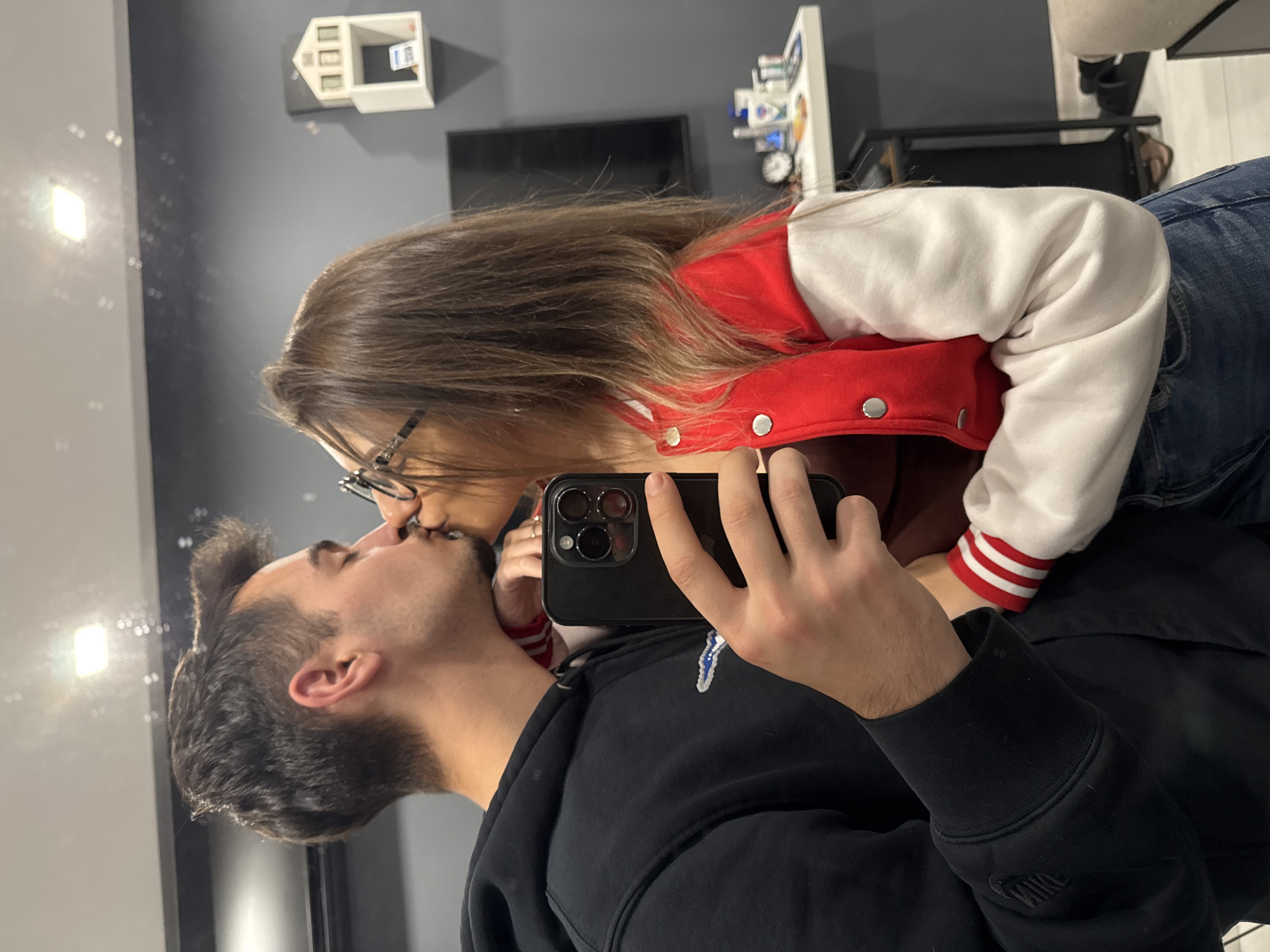 A couple is kissing while taking a selfie in front of a mirror. The woman is wearing glasses, a red and white jacket, and jeans. The man is wearing a black hoodie with a Nike logo on the sleeve. The background includes a grey wall with a small white house decoration and a black TV screen. There is also a white shelf with various items on it. The location is likely an apartment or home setting. The city of Sofia, Bulgaria cannot be determined from the image.