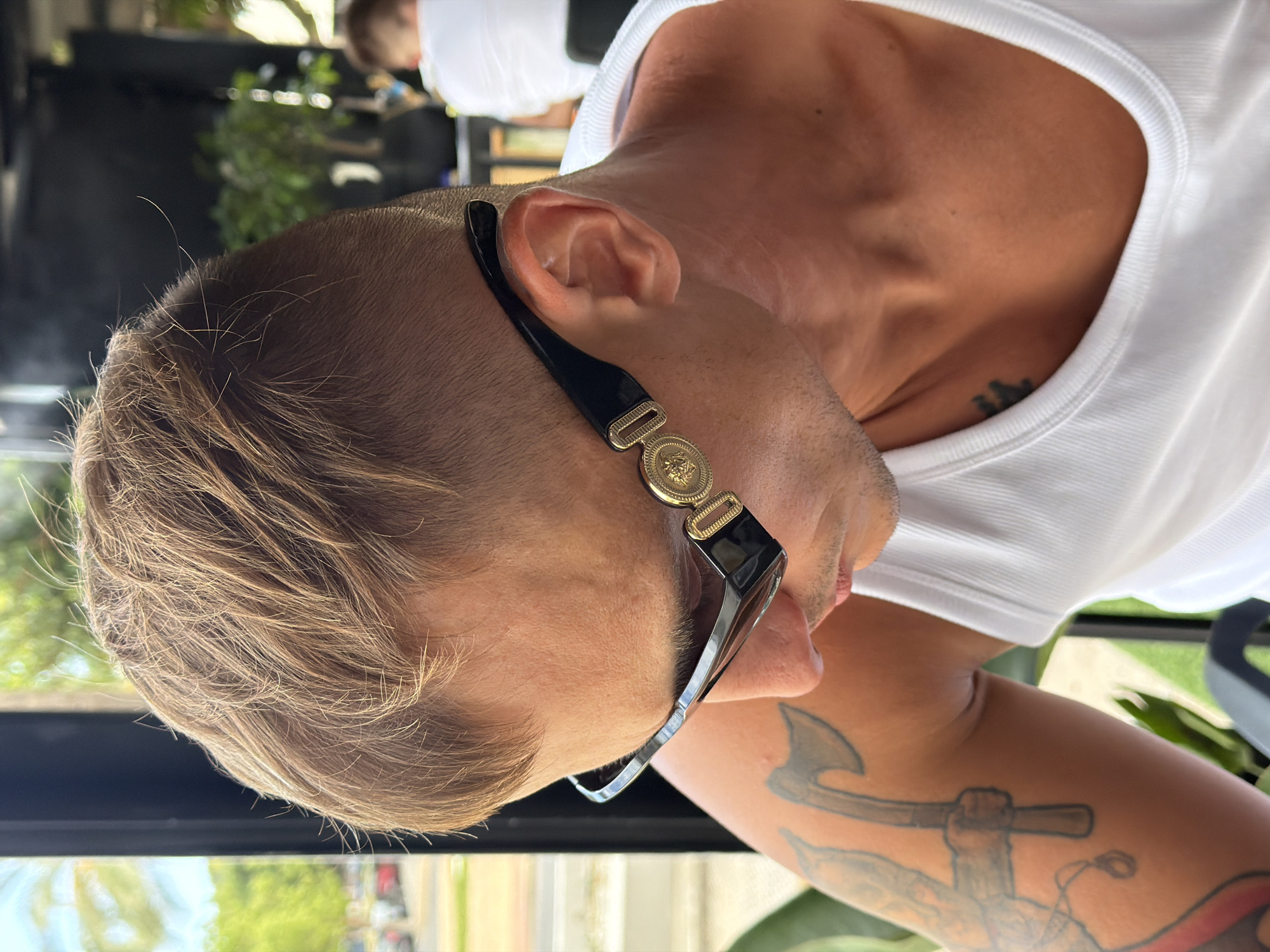 This is a close-up, rotated, vertical photograph of a person's head and shoulder, presumably outdoors in La Vila Joiosa, Spain. The subject is wearing a white tank top and large, fashionable sunglasses with black frames and a gold emblem on the temple. The sunglasses are positioned on their nose and the side of their face, obscuring the eye. The person has short, light brown hair and visible stubble on their chin and neck. There is a tattoo of an anchor on their left arm. In the background, blurred greenery suggests a lush, outdoor environment. The lighting indicates it is daytime, likely sunny. The overall impression is casual and stylish, with the focus on the person's accessories and facial profile.