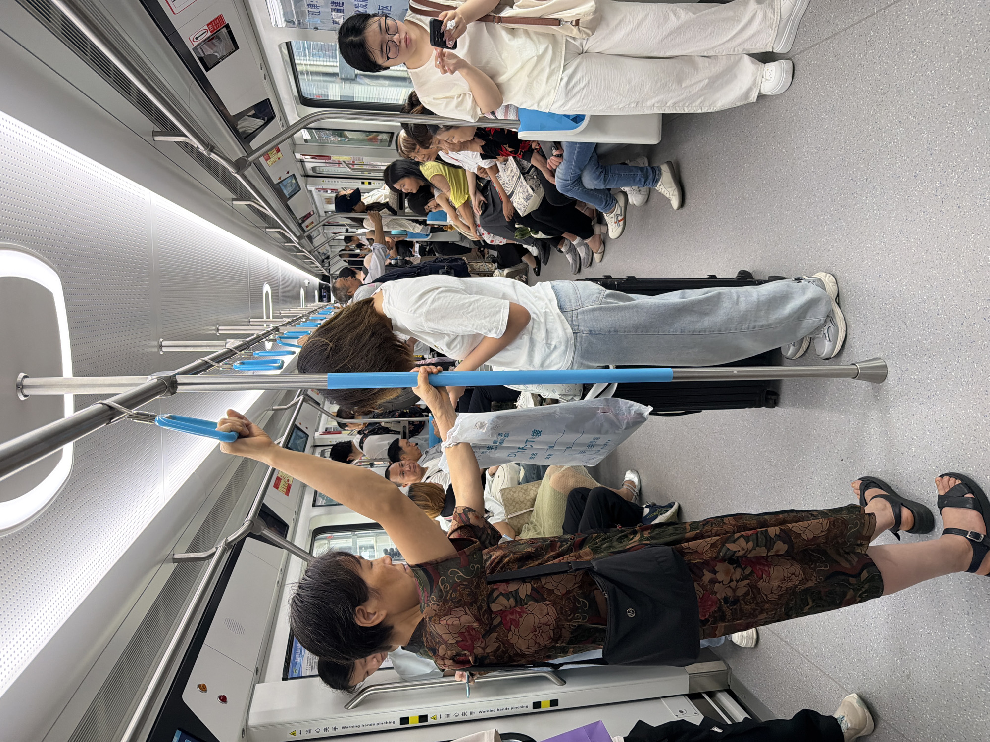 The image depicts the interior of a crowded metro train car, likely in Chongqing High-tech Zone, China, given the context. Several passengers are visible, some standing and holding onto overhead bars and poles, while others are seated. The overall atmosphere appears to be busy, typical of public transportation during peak hours.
In the foreground, a woman in a floral patterned dress is holding a white plastic bag and a blue vertical pole. She is looking downwards, and her left arm is raised, grasping a blue handrail. To her right, another woman in a white t-shirt and light blue jeans is also holding onto a pole. Her head is tilted downwards as she looks at her mobile phone.
Further into the car, a variety of individuals are observed. Some passengers are seated, including a woman in a yellow shirt and another in a black shirt with red accents. Others are standing, creating a densely packed environment. The train car itself is modern, with a white ceiling, metal poles, and grey flooring. Overhead lighting illuminates the space.
The scene captures a moment of everyday life on public transport, with individuals engaged in personal activities or simply enduring the commute. The image is a snapshot of urban life in a busy city, highlighting the shared experience of commuting.