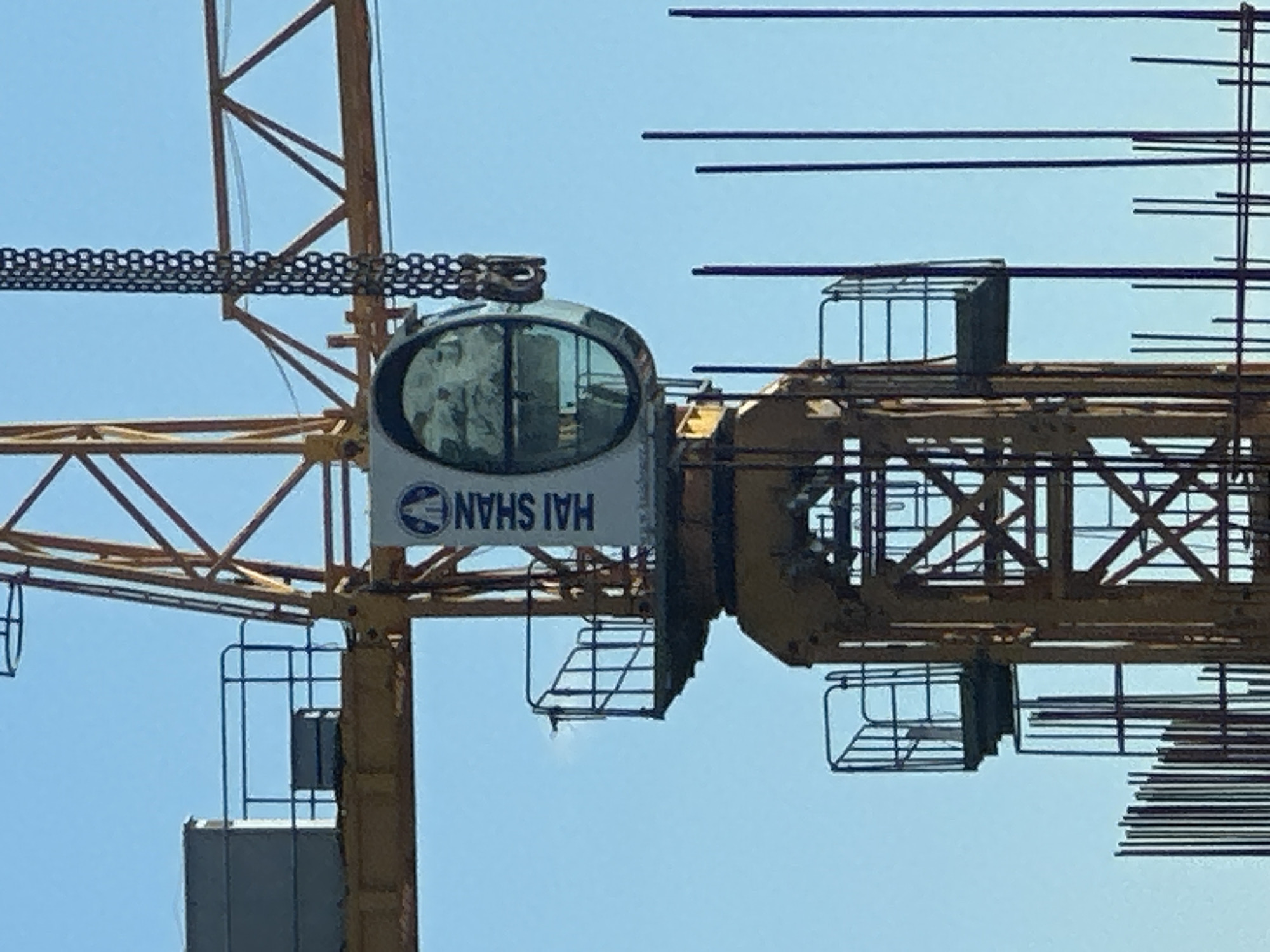 The image shows a construction crane against a clear blue sky. The crane is yellow and appears to be in operation. A portion of the crane's operator cabin is visible, which has a curved window and the words "HAI SHAN" printed vertically in blue text on a white background. A heavy chain hangs from the crane, with a hook at the end. Below the cabin, a section of the crane's tower structure is visible, which is made of interconnected yellow beams. In the foreground, vertical rebar rods are clustered together, indicating ongoing construction. The overall scene is consistent with a construction site in an urban environment. The image was taken in Kyiv, Ukraine.