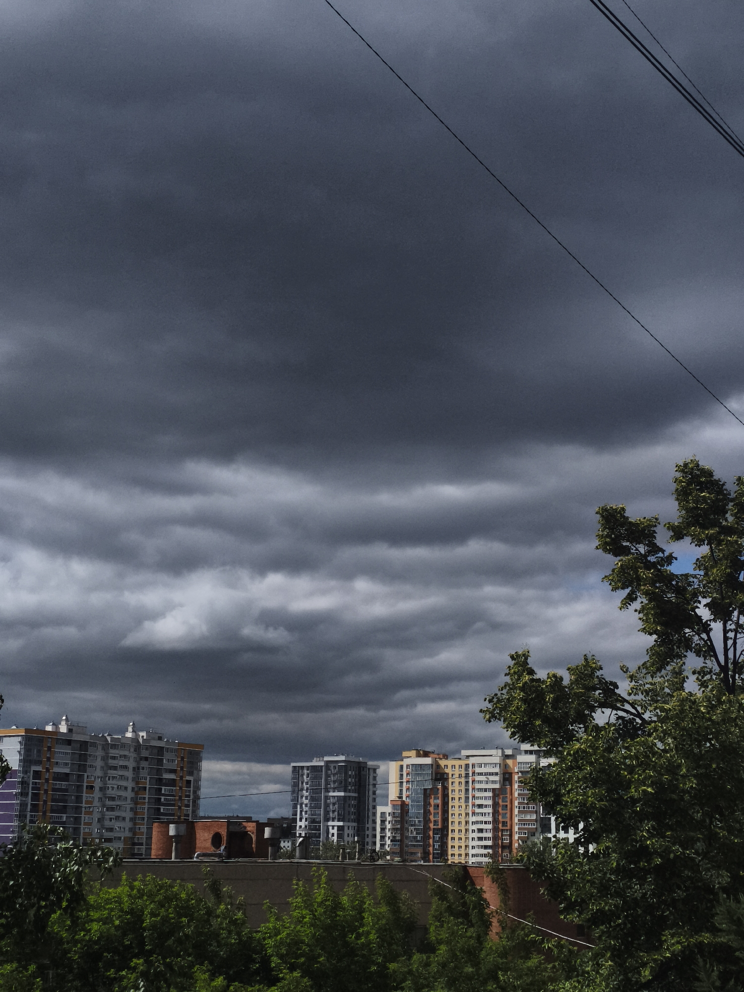 The image shows a view of apartment buildings in a city, with a stormy, dark sky overhead. There are several tall buildings visible, some with balconies and various colored facades. Trees with green foliage are in the foreground and partially obscuring the lower parts of the buildings. Power lines are visible in the upper right portion of the image. The overall mood is dark and overcast, suggesting impending rain or a storm. The specific location is not visually identifiable from the image alone.