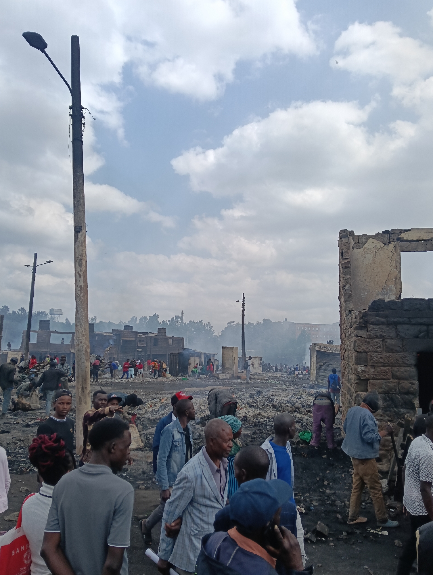 The image captures a scene of destruction in a densely populated urban area, likely a slum, in Nairobi, Kenya. The foreground is dominated by a utility pole, with visible wires and a street lamp attached. A crowd of people, appearing to be residents, are gathered in the aftermath of what seems to be a fire. Many of the structures in the background are either completely destroyed or heavily damaged, with some showing visible structural integrity loss and rubble. Smoke billows in the mid-ground, suggesting recent or ongoing burning. The ground is covered in debris, ash, and scattered personal belongings. People are observed walking through the wreckage, some looking around, others in conversation, and some seemingly searching through the debris. The atmosphere is somber, with a cloudy sky overhead. The presence of multiple damaged buildings and the large gathering of people point towards a significant incident such as a fire or demolition that has affected the community.