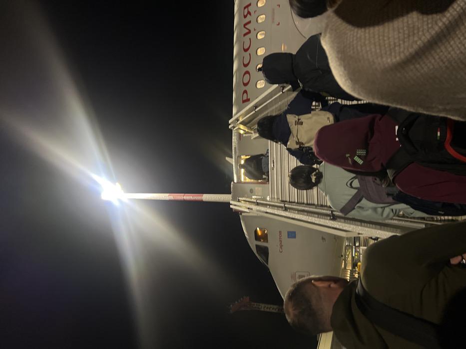 The image depicts a nighttime scene at Emelyanovo Airport, Russia, focusing on passengers boarding a commercial aircraft. A line of approximately seven to ten individuals, mostly viewed from behind, is ascending mobile boarding stairs to the aircraft's front-left entry door. The aircraft is a white narrow-body jet, featuring "РОССИЯ" (Russia) in red capital letters on its fuselage and "Саратов" (Saratov) below the cockpit windows. A bright, tall airport light pole with red and white striped sections stands to the left of the stairs, emitting a strong white glare that dominates the upper central portion of the frame. One person is partially visible entering the aircraft door, while others follow in succession. Distant airport infrastructure or a control tower with red and white markings is faintly visible in the far left background against the dark night sky. The primary light sources are the airport pole, the aircraft's interior, and external boarding area lighting.
