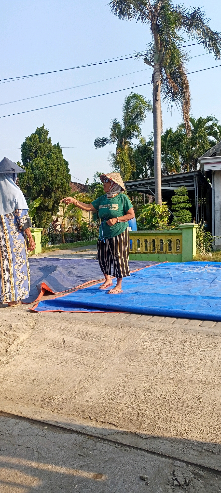 In Gempolklutuk, Indonesia, two individuals are observed outdoors. One person, viewed from the side and from behind, is wearing a light blue head covering and a long, patterned blue and gold robe, standing on the left side of the image. To their right, a person wearing a conical hat, sunglasses, and a green t-shirt with the text "BEAUTIFUL GIRL CLASSIC" printed on it, is actively gesturing with their right hand, index finger extended. This person is also wearing black and white striped pants that fall to mid-calf and light-colored flip-flops. They are standing on a large, unfolded blue tarp with orange trim, which is laid out on a concrete surface. In the background, palm trees, other foliage, a light-colored building, and a decorative green fence with yellow accents are visible under a clear sky. The lighting suggests it is daytime, likely morning or late afternoon due to the angle of the shadows.