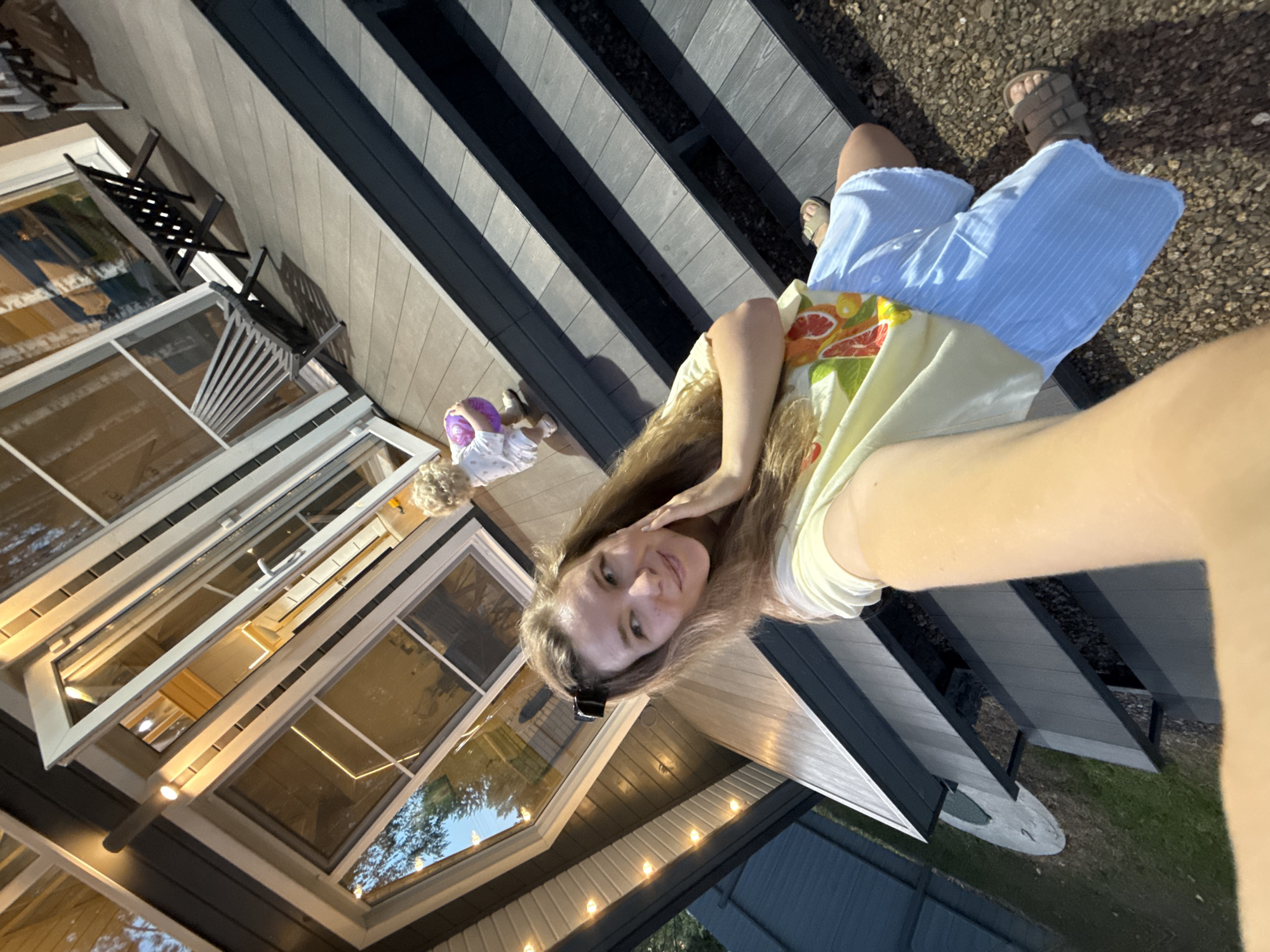 The image depicts a selfie of a young woman taken outdoors in what appears to be a residential setting. She is wearing a yellow t-shirt with a fruit print and light blue striped shorts, and sandals. Her hair is long and brown, pulled back with a black clip. She is touching her face with her hand and looking off to the side. In the background, a small child with blonde curly hair is visible, holding a purple ball. The child is wearing a white outfit. They are on a wooden deck or patio in front of a modern house with large windows. String lights are visible along the side of the house. The ground surrounding the deck is covered in gravel and grass. The overall impression is a casual, outdoor family scene. The image does not contain any specific landmarks or contextual clues that would allow for definitive identification of the location within Prokudino, Russia.
