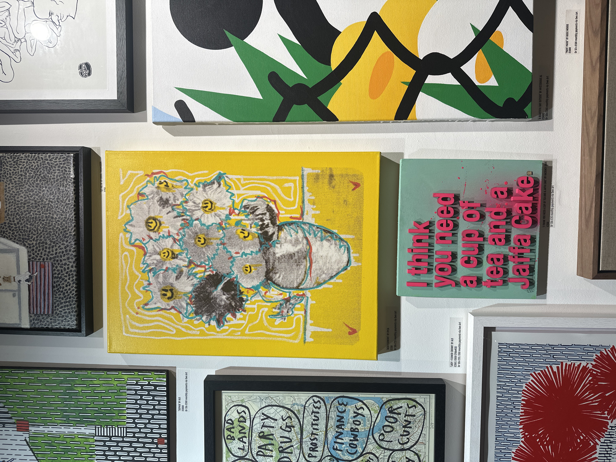 This media file captures a wall in an art gallery or exhibition, likely in London, United Kingdom, displaying a diverse collection of contemporary artworks. The scene shows multiple framed and unframed pieces hung closely together against a white wall, suggesting a curated display for viewing and sale.
The central focus is an unframed painting with a vibrant yellow background featuring stylized white and grey flowers, some with yellow smiley faces in their centers, and a vase-like object. This piece is titled "'ACID FLOWERS' BY RYCA" and priced at £150, or 10x £15 monthly payments via Own Art.
To its right, a mint green square canvas with raised pink 3D text reads: "I think you need a cup of tea and a Jaffa Cake". This artwork, by Hackney Dave, is priced at €250, or 10x €25 monthly payments via Own Art. Above the central yellow painting, a larger abstract piece, possibly titled "'A BEAUTIFUL DAY OUTSIDE BY MYSTERIOUS AL" (£2400) or "'PAINT PAUSE' BY ROSIE WOODS" (£1000), features bold black lines, yellow and green shapes, and white spaces.
Below the yellow floral piece, a framed artwork on a map-like background is visible, with hand-drawn bubbles containing words such as "BAD LANDS," "PARTY DRUGS," "PROSTITUTES," "FINANCE COWBOYS," and "POOR CUNTS". This piece is likely by ALO, with a label showing "'LADY + FLOWER CROWN' BY ALO £350 / £600 (FRAMED)".
Other visible artworks include a dark framed piece with a textured pattern and a small white building to the left, and a framed piece with green and white horizontal stripes, potentially by ALO, titled "'SOPHIA'" (£3000). On the far right, a framed artwork features red abstract shapes, possibly flowers, on a blue and white striped background. The consistent presence of price tags and "Own Art" payment plan details indicates that these works are offered for sale, typical of an art fair or gallery show. The lighting is bright and even, characteristic of an indoor exhibition space.