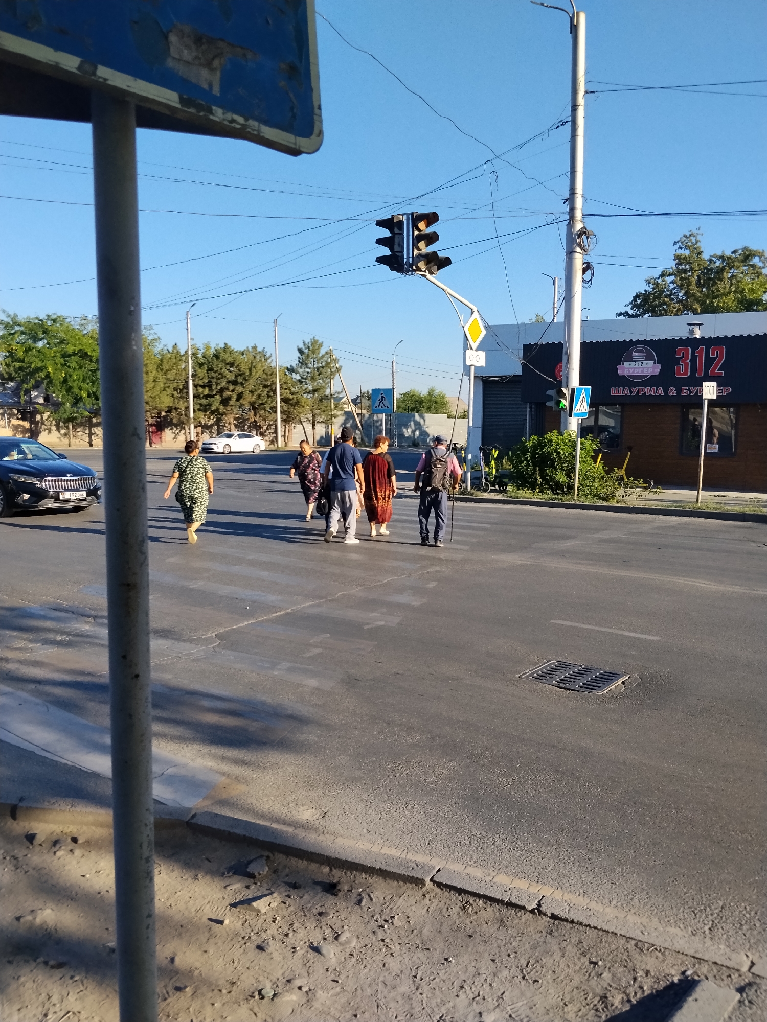The image depicts a street scene with several individuals crossing a pedestrian crossing. A black car is stopped on the left side of the road, while a white car is visible in the background. Traffic lights are illuminated, indicating a green signal. A building with signage that reads "312 SHURMA & BURGER" is situated in the background. The overall environment suggests an urban setting, possibly in Bishkek City, Kyrgyzstan, given the signage. The road surface appears to be asphalt, and there are streetlights and electrical wires visible. One person is wearing a floral print dress and walking away from the camera, while others are walking towards the camera or are in motion across the street. A person with a backpack and walking stick is also crossing. The sky is clear and blue, indicating fair weather.