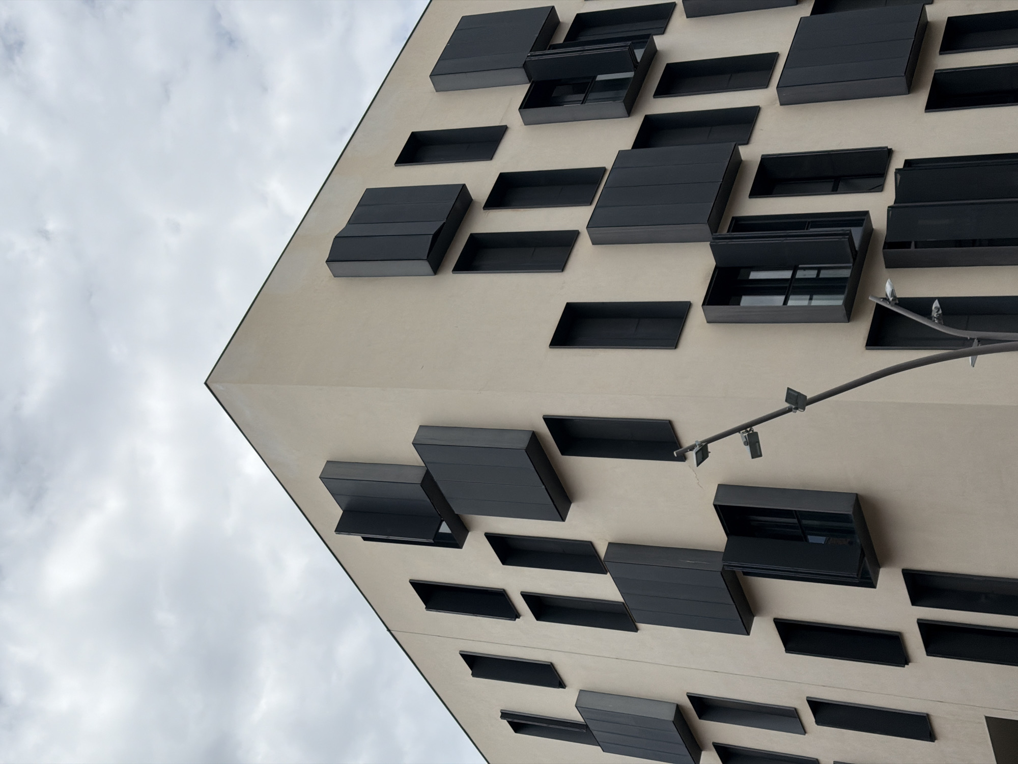 The image displays a modern building facade in Barcelona, Spain, captured from a low, upward-looking perspective against an overcast sky. The building features a light beige or off-white exterior wall that forms a sharp, angular corner diagonally across the left side of the frame. Integrated into this facade are numerous dark, almost black, rectangular and rhomboid-shaped elements. These elements vary in their orientation and depth; some appear flat against the wall, while others protrude significantly, functioning as either windows with deep frames or stylized sun shades. Several of these dark structures are partially open, revealing their shadowed interiors, which appear to house window panes or ventilation slats.
A thin, dark metal bar, possibly an exterior lighting track or a decorative architectural feature, runs horizontally then angles slightly upwards across the mid-right section of the building's surface, with small, light-colored fixtures attached to it. The sky, visible in the upper-left quadrant, is a uniform grey-white, indicating cloudy weather and diffused daylight. There are no people visible, and no discernible text appears on the building or in the scene. The overall impression is one of modern, minimalist architecture with strong geometric lines and a striking contrast between the light facade and dark window features.