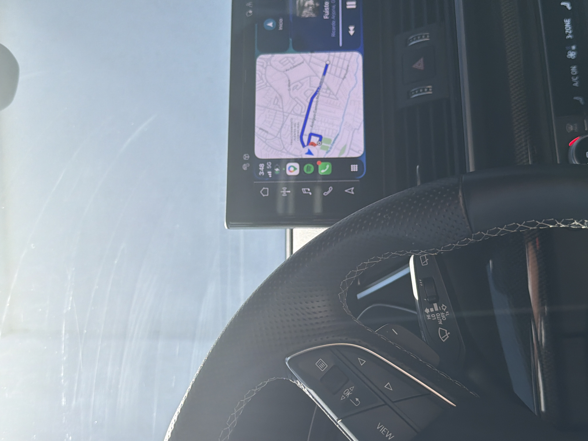 An interior shot of a car, likely from the driver's perspective, shows the steering wheel, dashboard, and an infotainment screen. The image is captured in bright daylight, with sunlight reflecting off the windshield, which appears to have some streaks or smudges. No people are visible.
The black leather steering wheel, with distinctive white stitching and a perforated grip, occupies the lower right portion of the frame. It features multifunction buttons on the left spoke, including one labeled "VIEW" and other controls. To the right of the steering wheel, a wiper control stalk is visible, with settings like "HI", "LO", "AUTO", and "OFF" printed on it.
A prominent infotainment screen is mounted centrally on the dashboard, displaying a car-specific interface. The screen shows the time as "3:48" and a "5G" network connection. A navigation map, likely depicting a route in San Bernardino Tlaxcalancingo, Mexico (with visible text "Antiguo Camino Real"), is centrally displayed within a card. Below the map, several app icons are visible, including Google, Spotify, and a phone icon. Above the map, a music player displays "Fuiste" by "Ricardo Arjona".
The dashboard features horizontal air vents and a prominent red triangular hazard light button. To the right of the screen, parts of the climate control panel are visible, indicating "3-ZONE" climate control and "A/C ON." The overall setting suggests the vehicle is either parked or being driven.