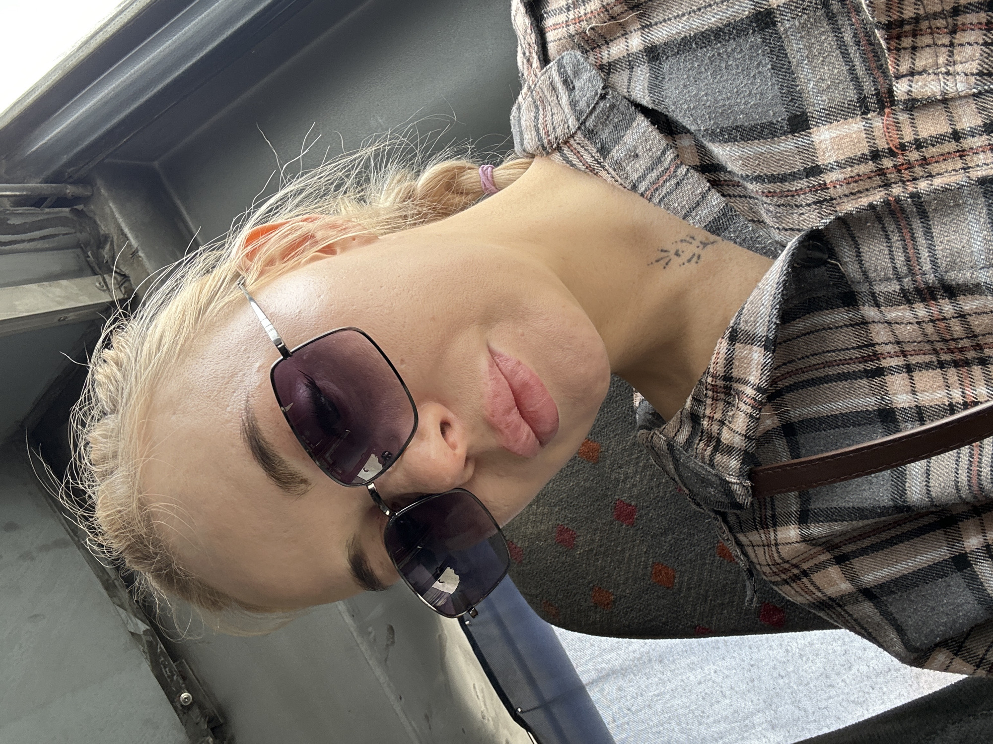 A young woman with blonde hair partially tied back, wearing sunglasses and a gray and brown plaid shirt over a gray shirt with red polka dots, is positioned in what appears to be a bus or train. The background suggests a public transportation setting with metallic structures and gray surfaces. No information confirms the location as Megion, Russia.