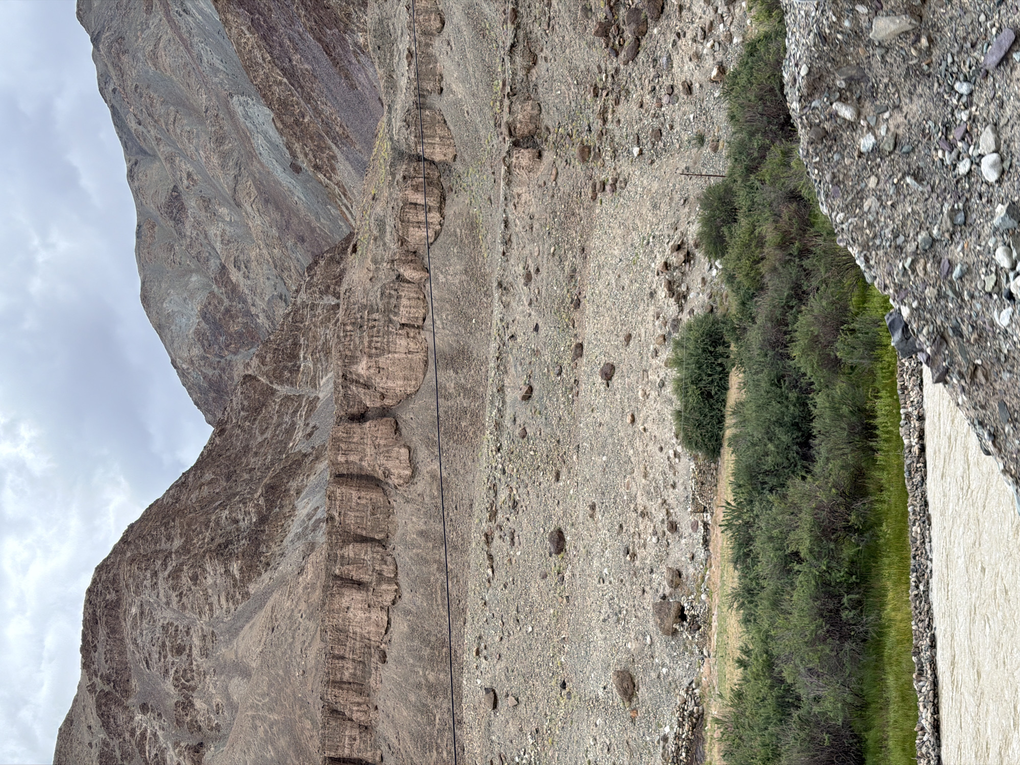 The image shows a wide, muddy river flowing in the foreground. On the right side of the river, there is a pile of grey and brown rocks and gravel. Beyond the river and rocks, there are green bushes and grasses growing. In the background, a vast, barren landscape of rocky mountains under a cloudy sky dominates the scene. The mountains are a mix of brown and grey, with layered rock formations and sparse vegetation. A thin black wire stretches horizontally across the middle of the frame, running parallel to a distinct ledge of rocky outcroppings. Some small, stone structures are visible in the middle ground, nestled amongst the vegetation. The overall impression is a remote, rugged natural environment. This scene is consistent with the landscape found in Kiari, India, known for its mountainous terrain and rivers.