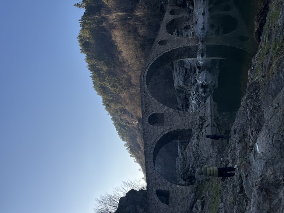 The image displays a multi-arch stone bridge spanning a river in the city of Diadovtsi, Bulgaria. The bridge, constructed of large grey stones, features at least six visible arches, three of which are prominent in the central frame. The river below has calm, reflective dark water, with rocky banks and exposed boulders visible in and around the water. On the left riverbank in the foreground, two individuals are present. One person, positioned on the left, wears a hooded, two-tone jacket (olive green on top, lighter green/yellowish on bottom), dark pants, and white sneakers, standing with their back to the viewer and facing the bridge. A second individual, dressed in dark clothing, stands further to the right and slightly closer to the water's edge, also facing the bridge. The background consists of a steep hillside on the right, densely covered with green evergreen trees, transitioning to sparser bare deciduous trees and rocky outcrops towards the left. A clear, bright blue sky is visible above the hills. Sunlight appears to originate from the left, illuminating the upper parts of the landscape and the bridge while casting shadows on the lower river area.