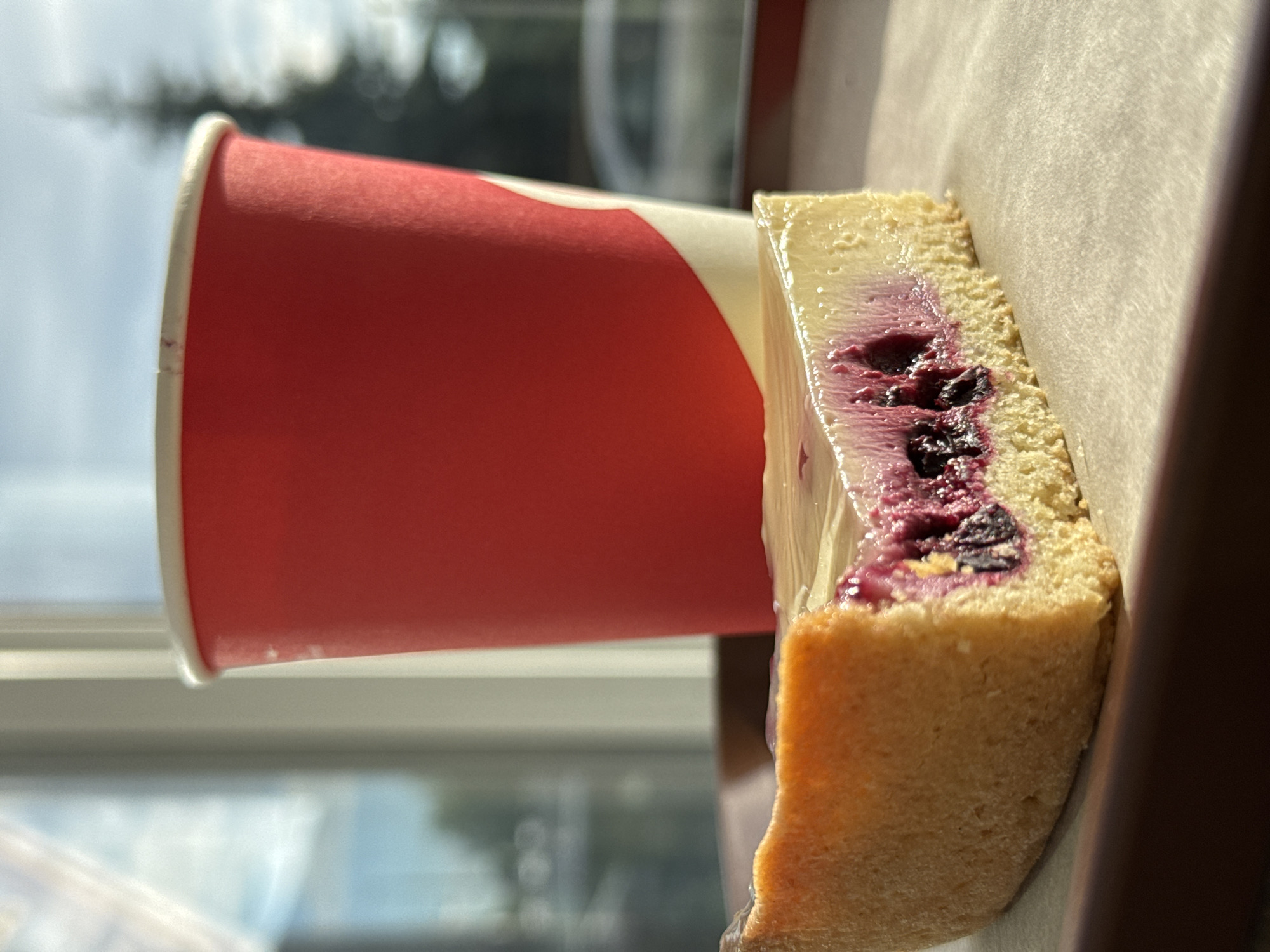 A close-up, vertical shot shows a slice of cheesecake with a berry swirl next to a red paper cup. The cheesecake has a light brown crust and a creamy white filling with deep purple berries visible throughout. The cup is plain red with a white rim. Both items are resting on a light brown surface, possibly parchment paper, which is partially illuminated by sunlight casting shadows. In the background, out of focus, are reflections of a window with sky and trees, suggesting an indoor setting near a window. The lighting indicates it is daytime, with the sun creating a warm glow on the cheesecake. There are no people visible, and the image focuses on the food and drink. There is no visible text on the cup or elsewhere in the image. The overall mood is casual and inviting, possibly capturing a moment of enjoying a treat.