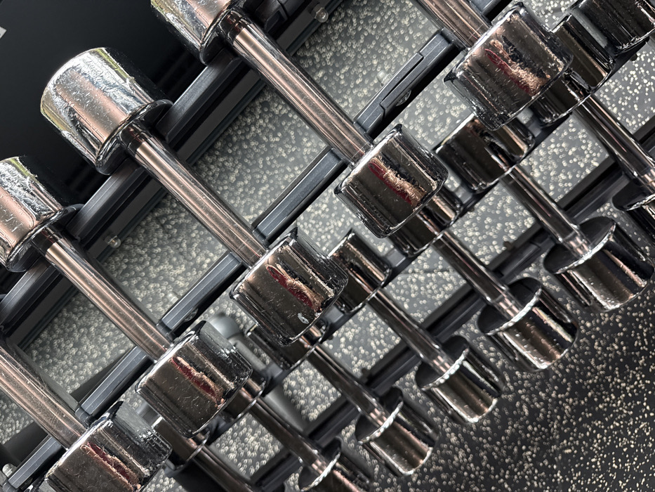 A row of chromed dumbbells is depicted, arranged diagonally upwards from left to right across the frame. The dumbbells feature polished metallic handles and cylindrical weight heads, resting securely on a dark, multi-tiered rack structure. Minor scuffs and surface imperfections are visible on the reflective chrome surfaces. Indistinct reflections are present on the dumbbell heads, showing blurred shapes and light sources, but no identifiable objects or individuals. The floor beneath the rack is a dark, speckled rubber material. The background beyond the rack is uniformly dark and out of focus. No persons are present, and no activities are taking place. The visual evidence within the image does not provide sufficient information to specify its location within the City of London, United Kingdom.
