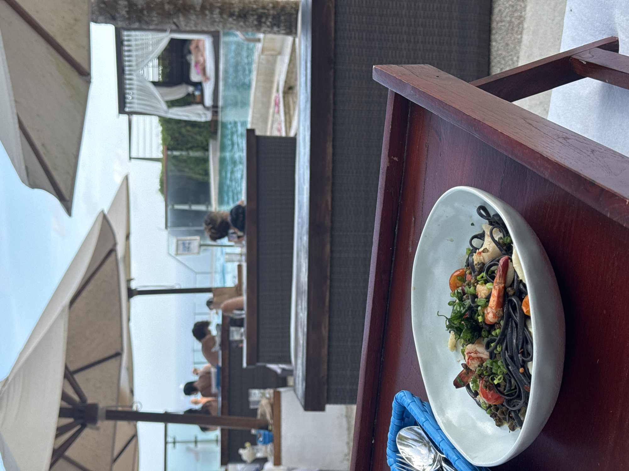 Here's a description of the image:
The scene appears to be at a resort or high-end hotel in an unknown city. In the foreground, a wooden tray holds a white bowl of squid ink pasta with shrimp and vegetables. A blue basket containing silverware sits alongside the bowl. In the background, a multi-tiered pool area is visible, with sun umbrellas and people lounging at the pool edge. There is also some resort-like furniture, such as beds and covered seating areas, closer to the pool.