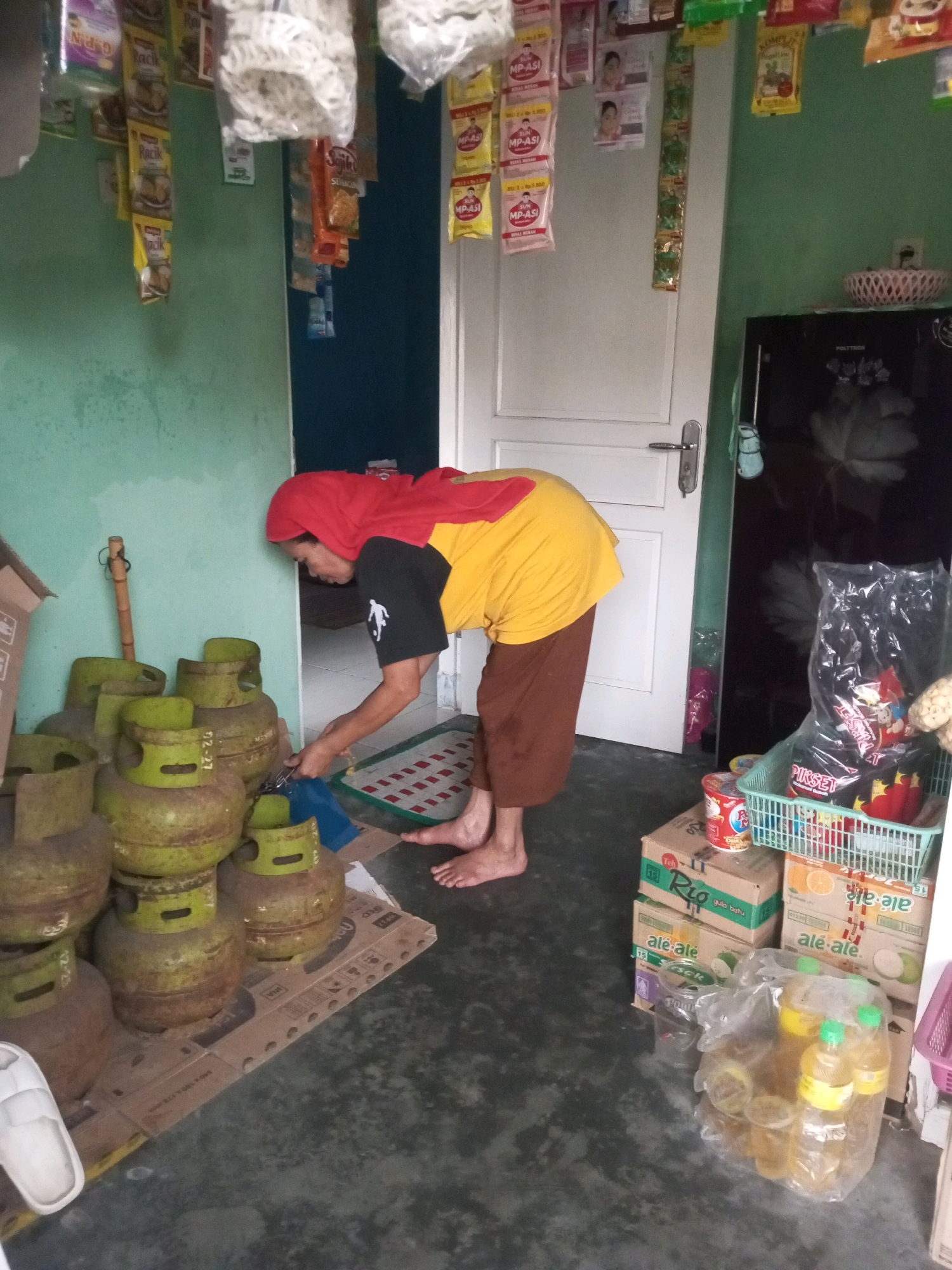 The image depicts an indoor scene within a small shop, possibly a warung, located in Andongsili, Indonesia. A person wearing a red headscarf and a yellow shirt over a black t-shirt is bending down, interacting with a stack of green gas cylinders. The person is barefoot. In the background, a white door is visible, and to the right, a black refrigerator with items on top and a basket containing snacks and drinks. Various packaged goods hang from the ceiling and walls, suggesting a retail environment. Cardboard boxes and plastic containers with beverages are also present on the floor. The overall impression is of a cluttered, functional space, typical of a local convenience store in Indonesia.