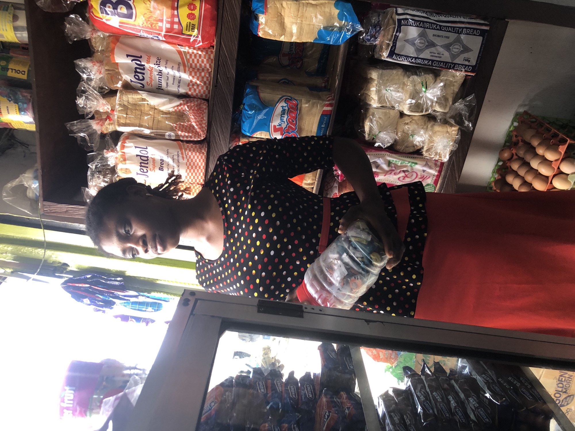 A young Black woman stands in a small shop, possibly in Ado, Nigeria. She is wearing a black, polka-dotted top with red trim and a long, bright red skirt. Her dark hair is pulled back, and she is holding a large, clear plastic bottle filled with various items. Her gaze is directed upwards and to the left.
The shop shelves behind her are stocked with numerous food items. On the top shelf, yellow and red packaged goods are visible. The middle shelves are filled with loaves of bread in clear plastic packaging, some labeled "Jendol Superb Bread" and "Skymart Bread." The lower shelves also contain bread and what appear to be bags of snacks or other packaged goods. To the left of the woman, a refrigerated display case shows stacks of dark-colored packaged items, some with "M'vites DARK" printed on them. Below the shelves, a carton of brown eggs is arranged. The lighting in the shop is bright, with natural light streaming in from the left, creating some glare on the display case.